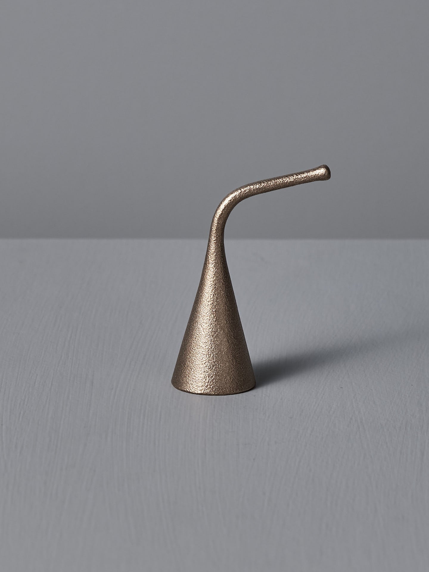 A Sammu Candle Snuffer by Nedre Foss, featuring a brass-colored, conical design with a curved handle, is placed on a gray surface against a light gray background, evoking traditional Finnish design.