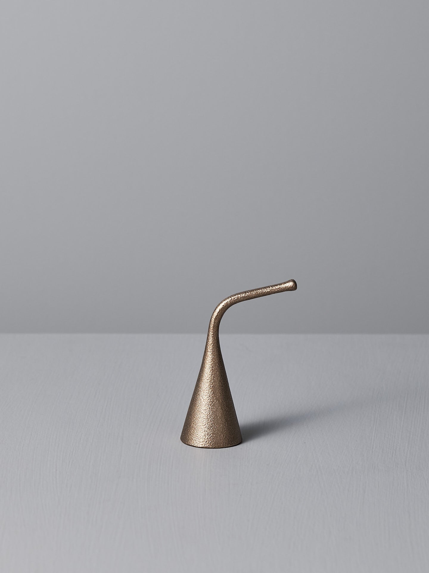 A Sammu Candle Snuffer by Nedre Foss, featuring a brass-colored, conical design with a curved handle, is placed on a gray surface against a light gray background, evoking traditional Finnish design.