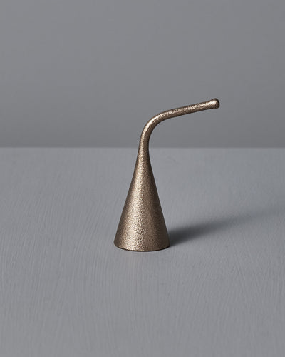 A Sammu Candle Snuffer by Nedre Foss, featuring a brass-colored, conical design with a curved handle, is placed on a gray surface against a light gray background, evoking traditional Finnish design.