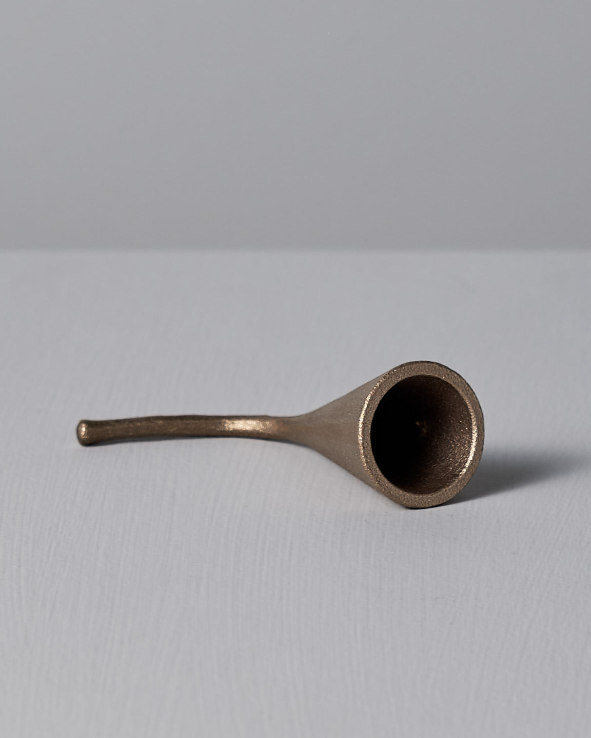 A Sammu Candle Snuffer by Nedre Foss, featuring a brass-colored, conical design with a curved handle, is placed on a gray surface against a light gray background, evoking traditional Finnish design.