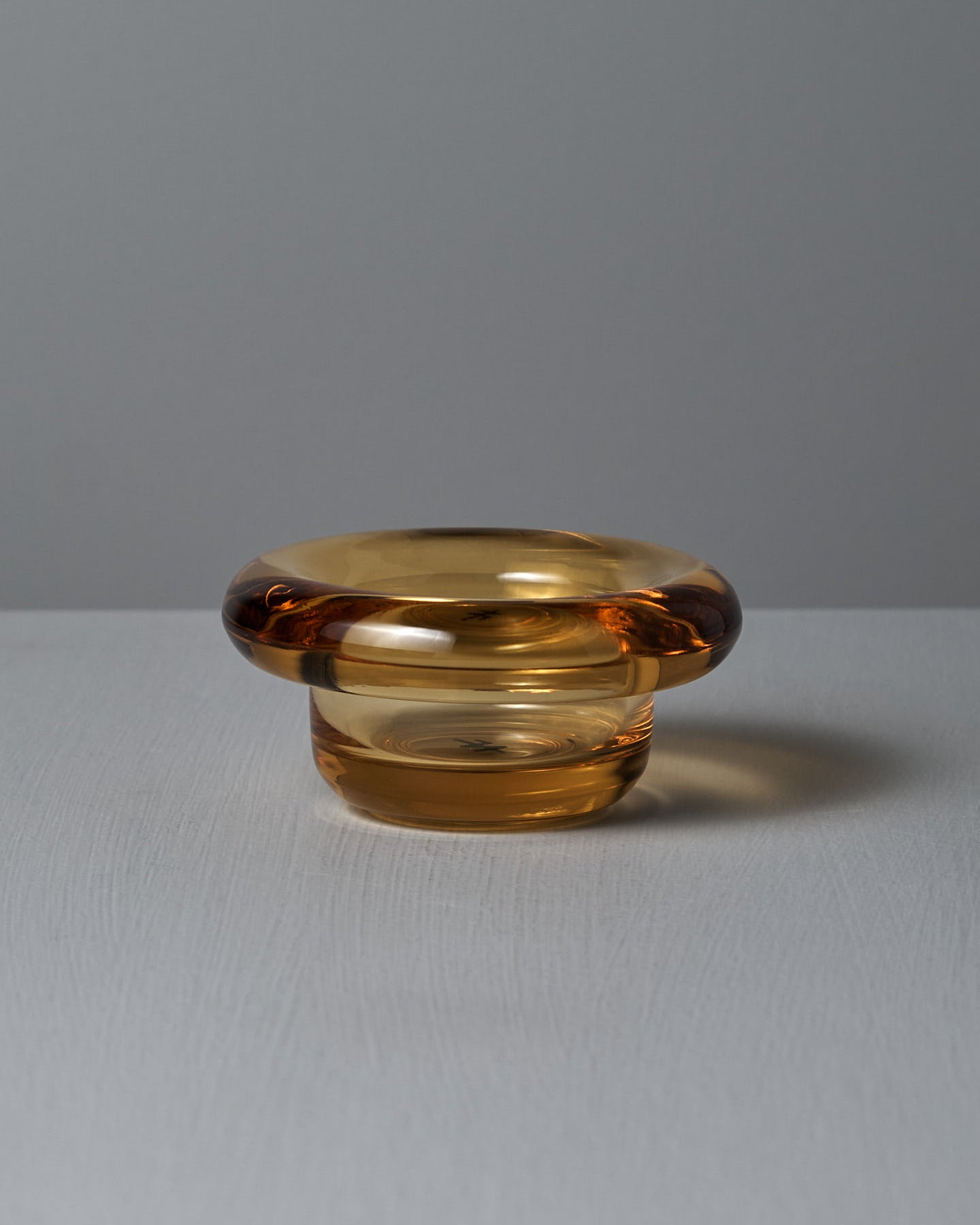 On a textured light gray surface, the Sirkel Candle Holder – Amber Yellow from Nedre Foss sits elegantly as a tea-light holder, its small transparent glass with a wide rim standing out against the plain gray background.