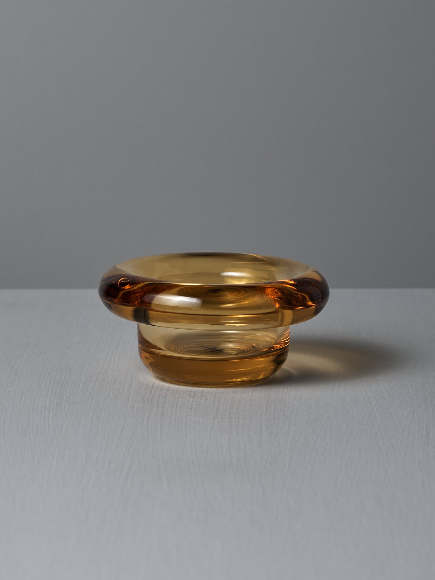 On a textured light gray surface, the Sirkel Candle Holder – Amber Yellow from Nedre Foss sits elegantly as a tea-light holder, its small transparent glass with a wide rim standing out against the plain gray background.