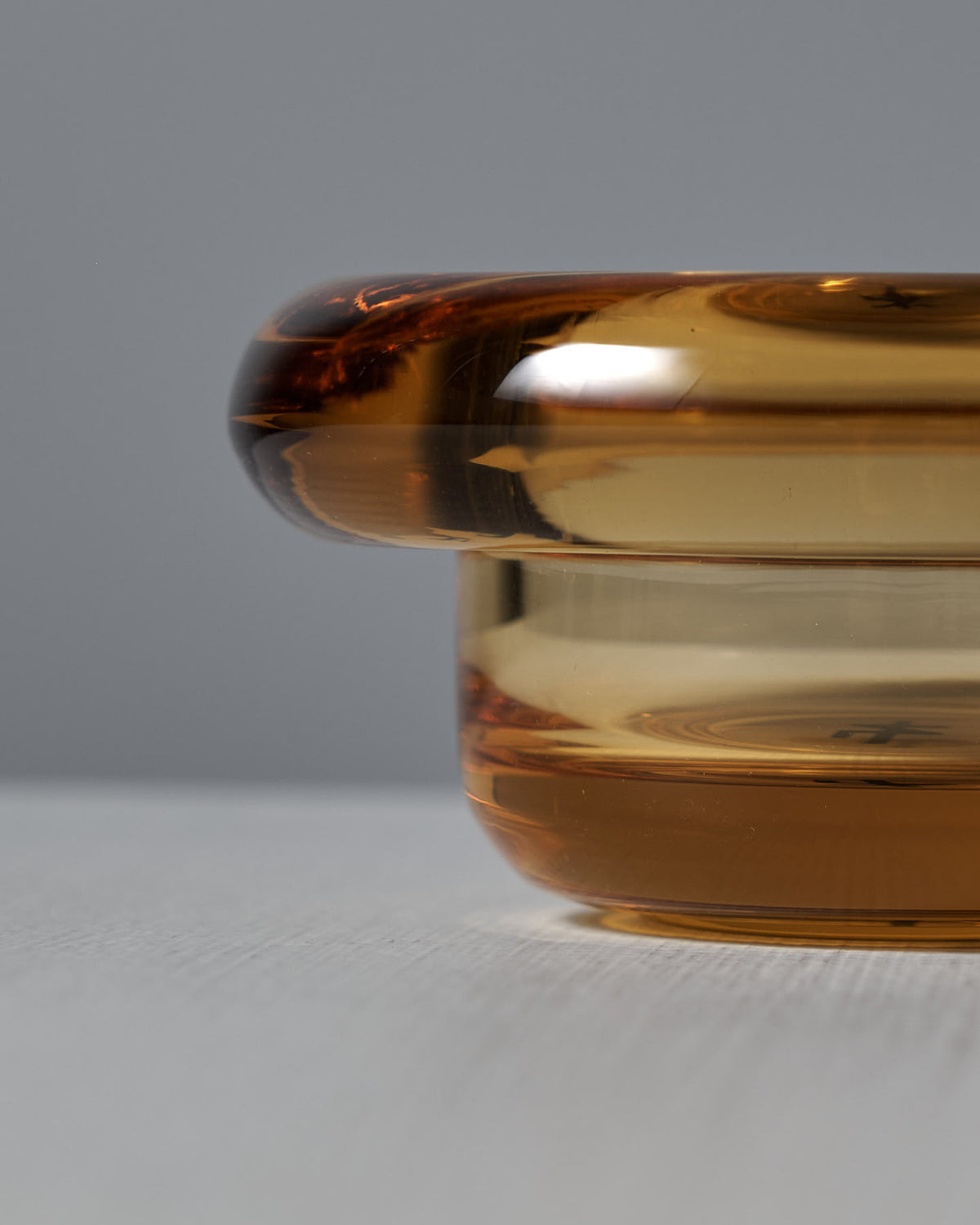 Close-up of the Sirkel Candle Holder – Amber Yellow by Nedre Foss, made of transparent amber glass with a rounded top edge, placed on a light surface against a gray background.