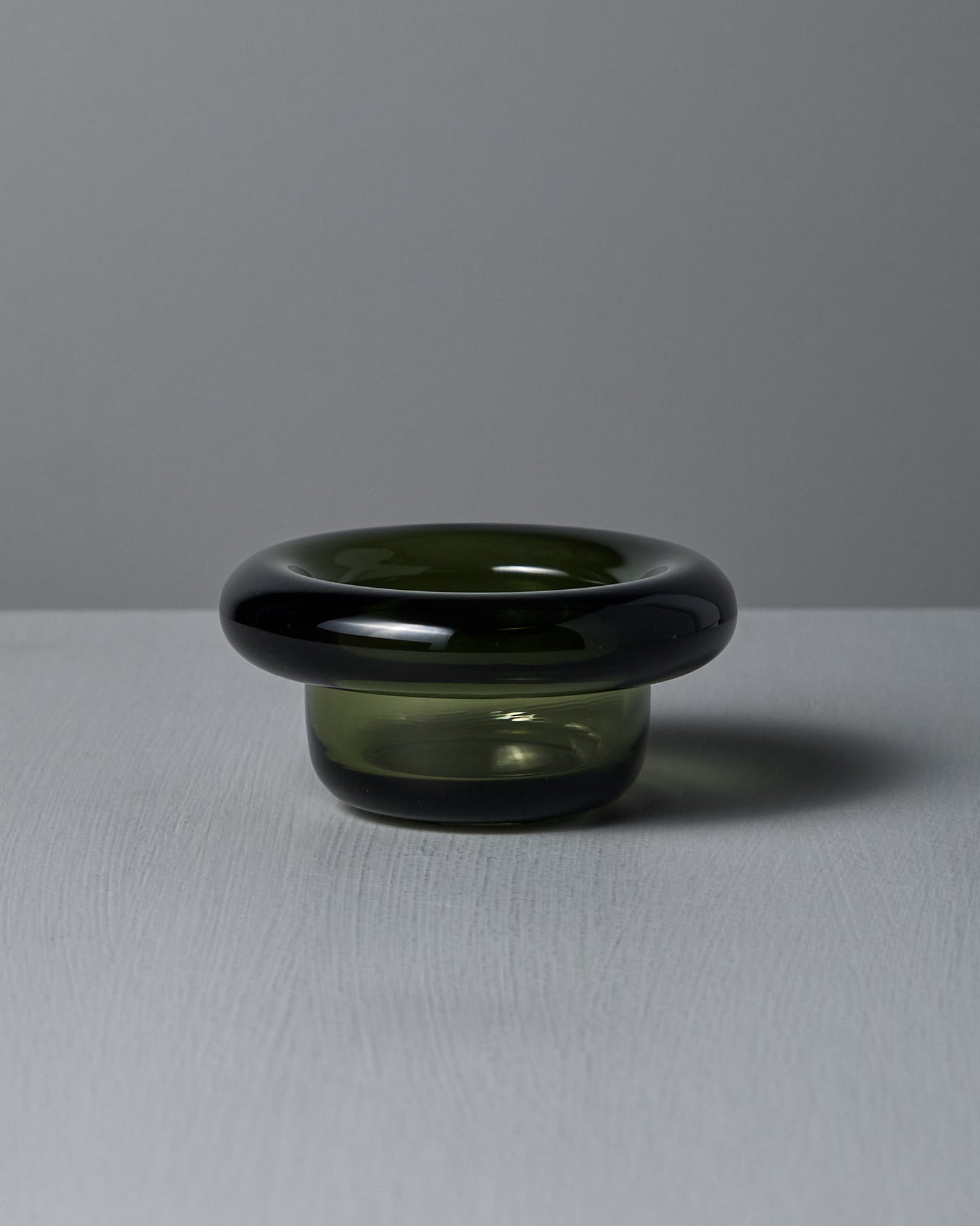The Sirkel Candle Holder – Forest Green by Nedre Foss, a pressed glass piece with a wide round rim and short base, rests elegantly as a tea-light holder on a textured gray surface.