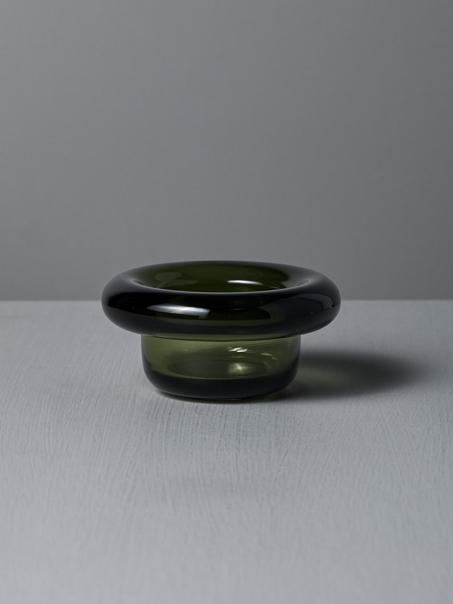 The Sirkel Candle Holder – Forest Green by Nedre Foss, a pressed glass piece with a wide round rim and short base, rests elegantly as a tea-light holder on a textured gray surface.