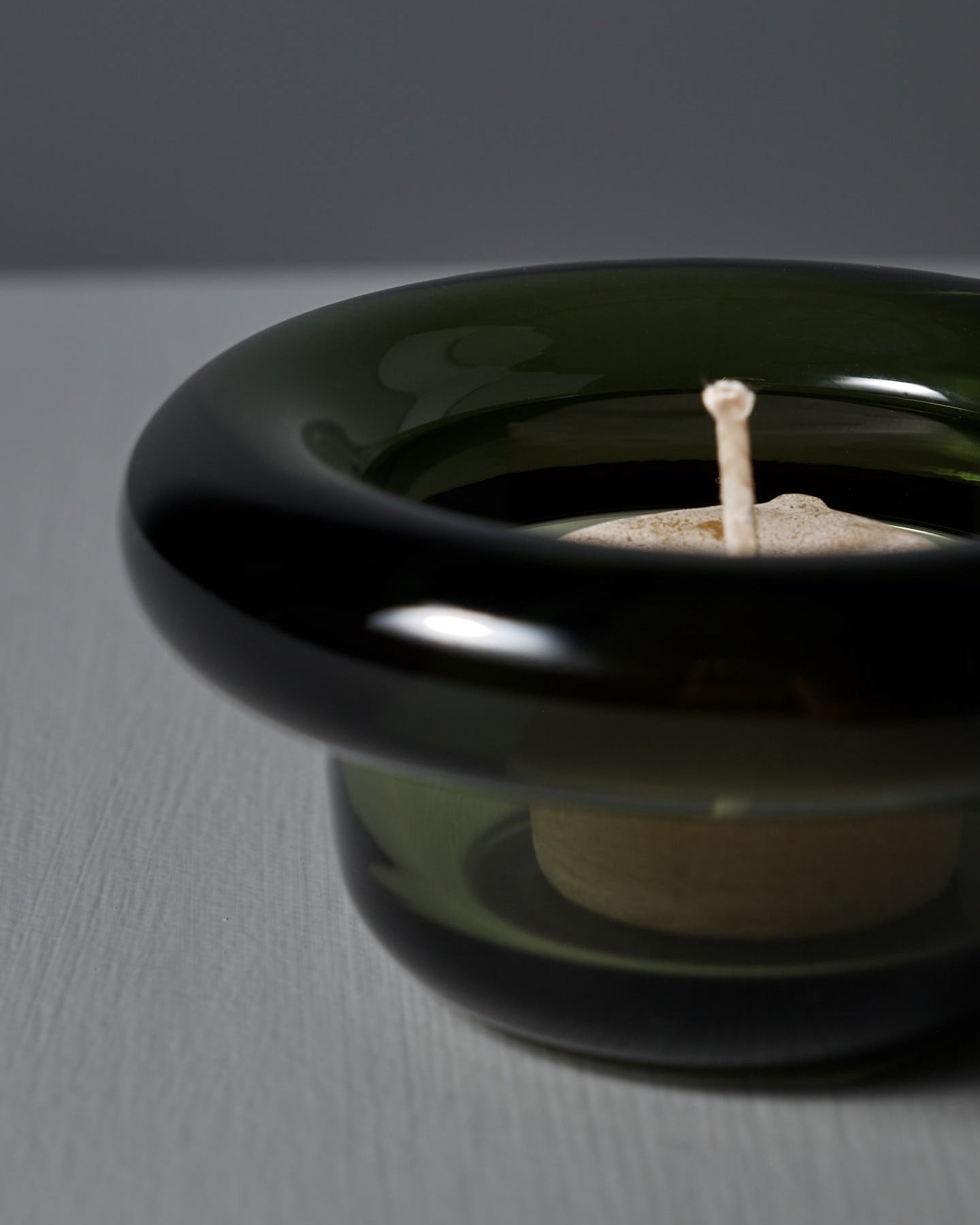 The Sirkel Candle Holder – Forest Green by Nedre Foss, a pressed glass piece with a wide round rim and short base, rests elegantly as a tea-light holder on a textured gray surface.