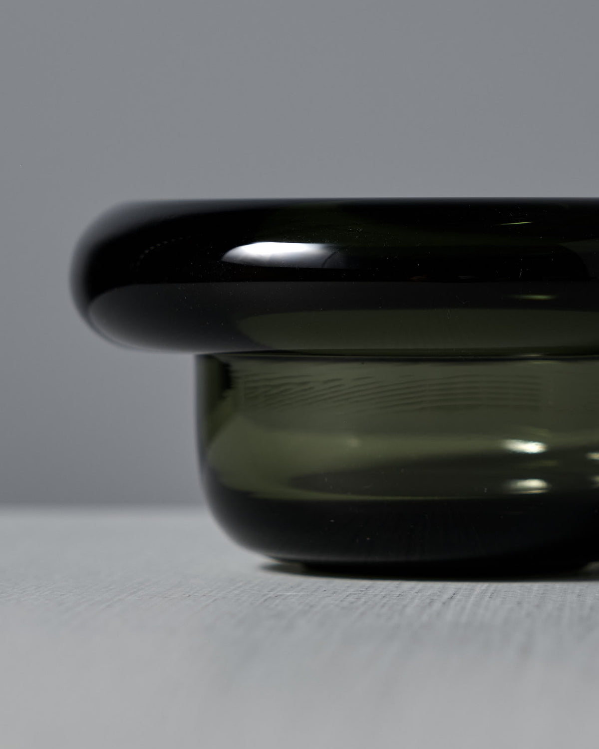 A close-up of the Sirkel Candle Holder – Forest Green by Nedre Foss, a glossy dark green pressed glass bowl with a wide rim, placed on a textured gray surface against a plain gray background.