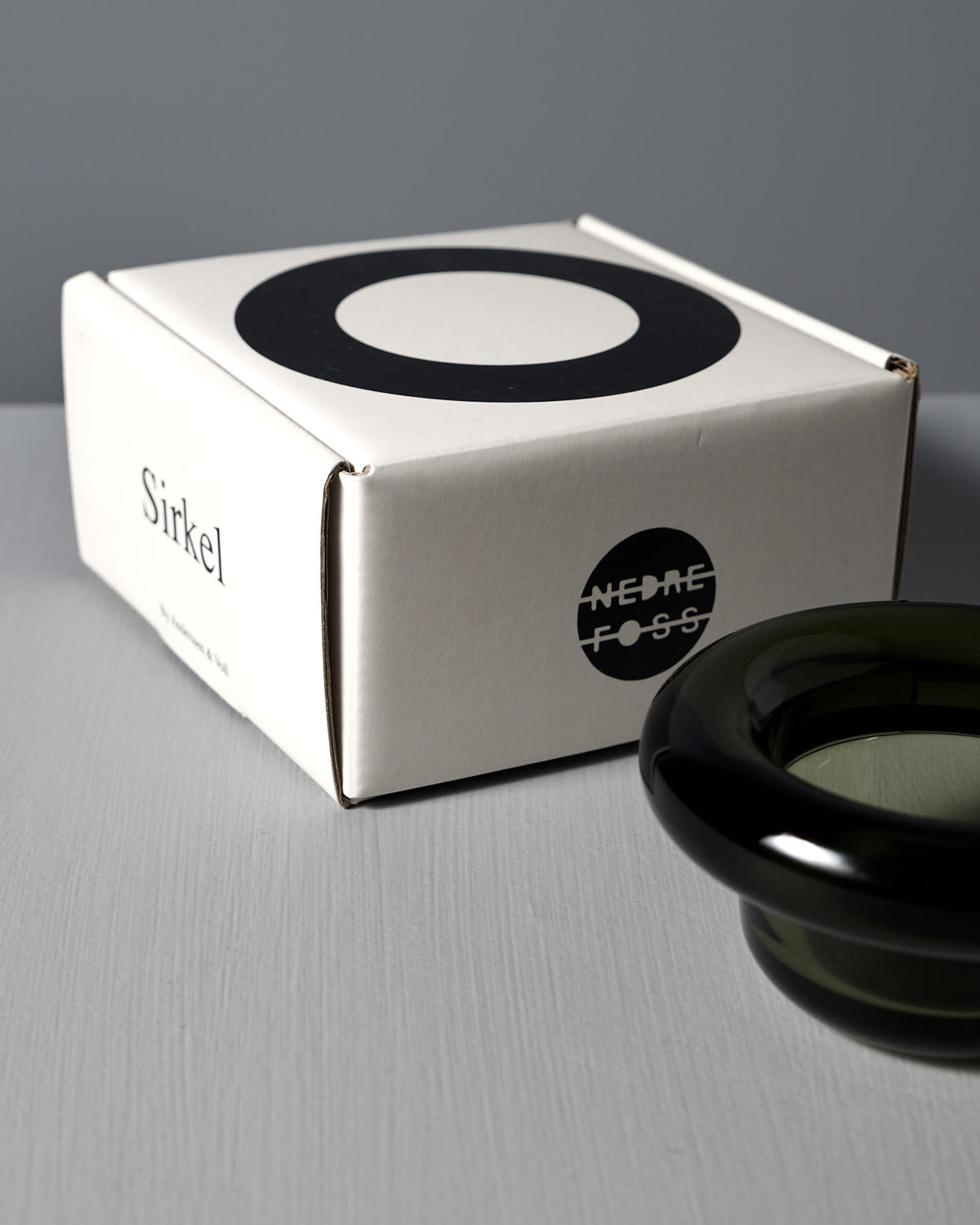 A white box labeled "Sirkel" is on a gray surface, next to the Nedre Foss Sirkel Candle Holder in forest green pressed glass.