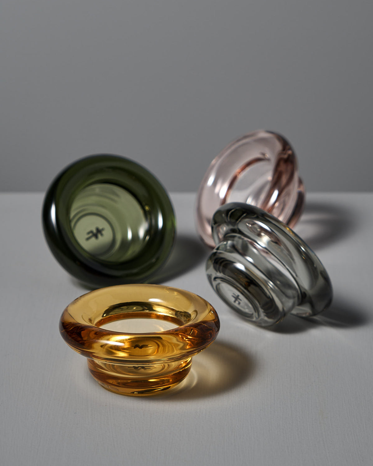Four elegant Sirkel pieces in green, primrose pink, gray, and amber glass from Nedre Foss are arranged on a flat surface. These rounded objects resemble tea-light holders and exhibit the artistry of pressed glass craftsmanship.