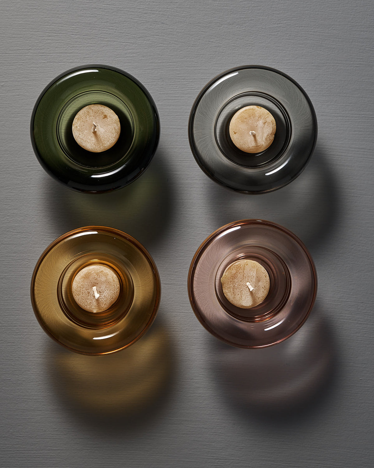 Four Nedre Foss Sirkel Candle Holders made of pressed glass sit on a gray surface, each holding a small candle. The round holders come in vibrant colors—amber yellow, black, green, and pink—casting elegant shadows to the right.
