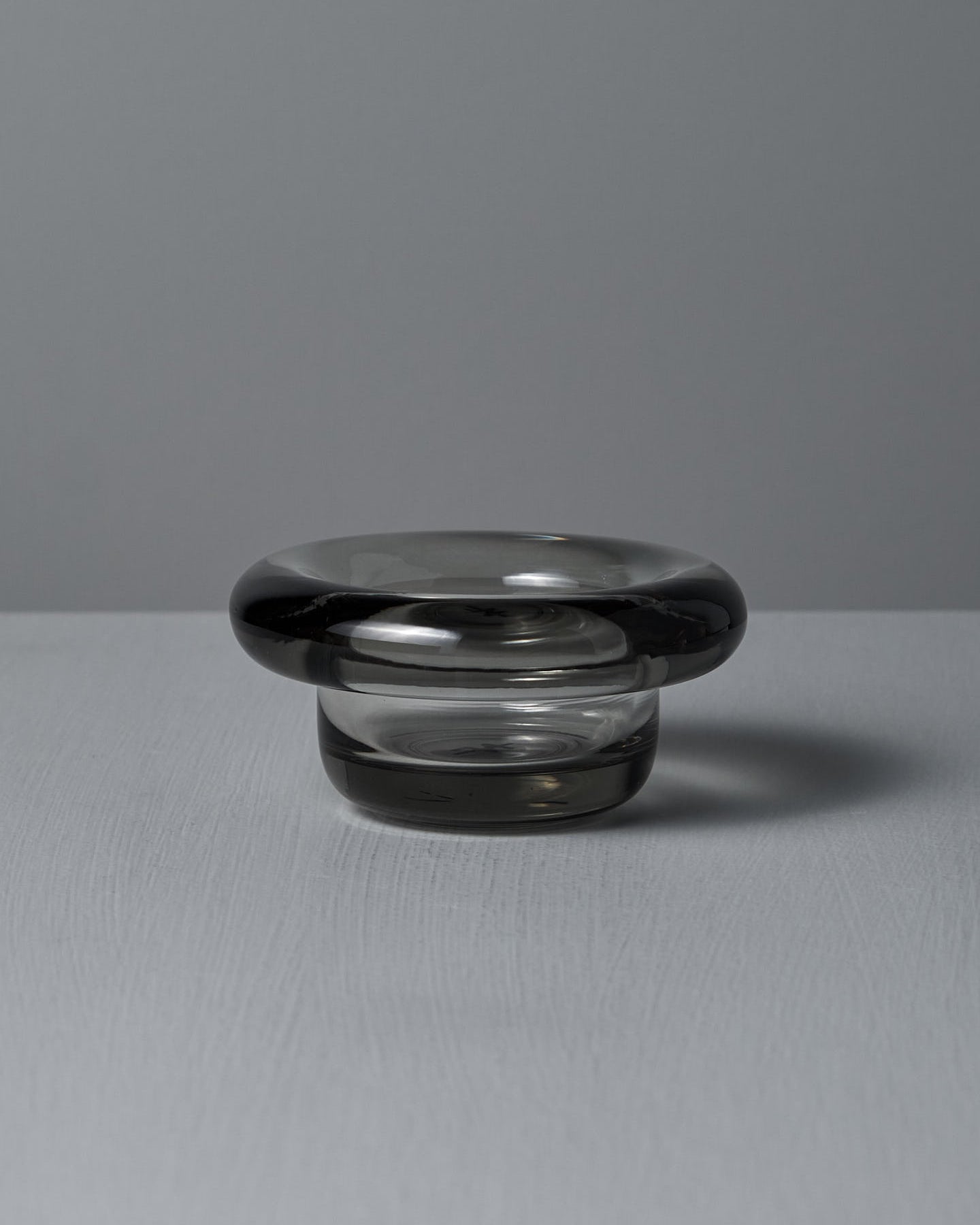 The Sirkel Candle Holder in translucent dark pressed glass by Nedre Foss features a wide, rounded rim and is elegantly suited as a tea-light holder on the smooth gray surface, enhancing the minimalist aesthetic.