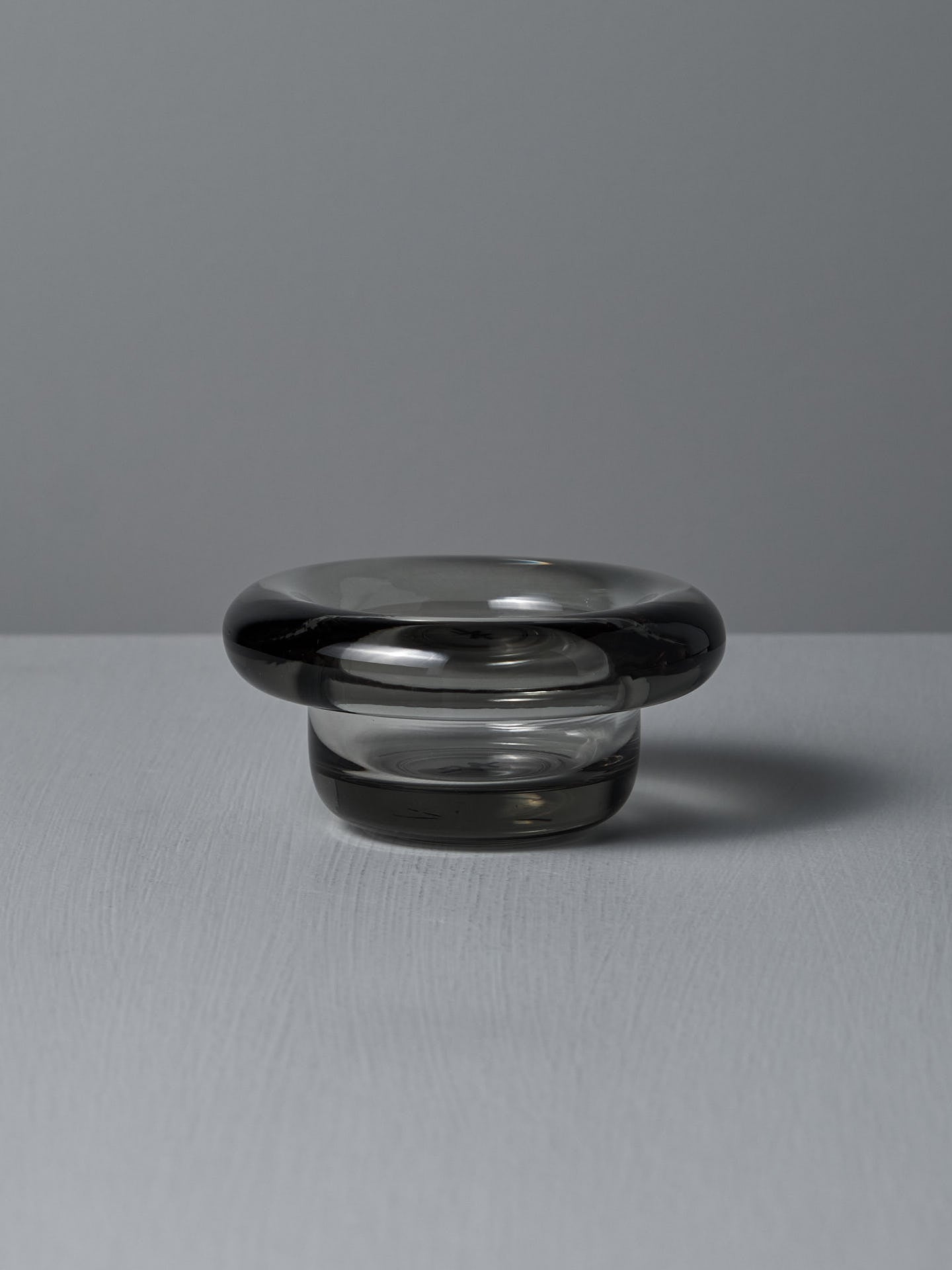 The Sirkel Candle Holder in translucent dark pressed glass by Nedre Foss features a wide, rounded rim and is elegantly suited as a tea-light holder on the smooth gray surface, enhancing the minimalist aesthetic.