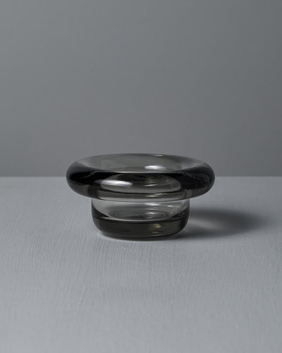 The Sirkel Candle Holder in translucent dark pressed glass by Nedre Foss features a wide, rounded rim and is elegantly suited as a tea-light holder on the smooth gray surface, enhancing the minimalist aesthetic.
