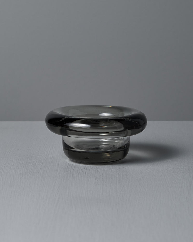 The Sirkel Candle Holder in translucent dark pressed glass by Nedre Foss features a wide, rounded rim and is elegantly suited as a tea-light holder on the smooth gray surface, enhancing the minimalist aesthetic.