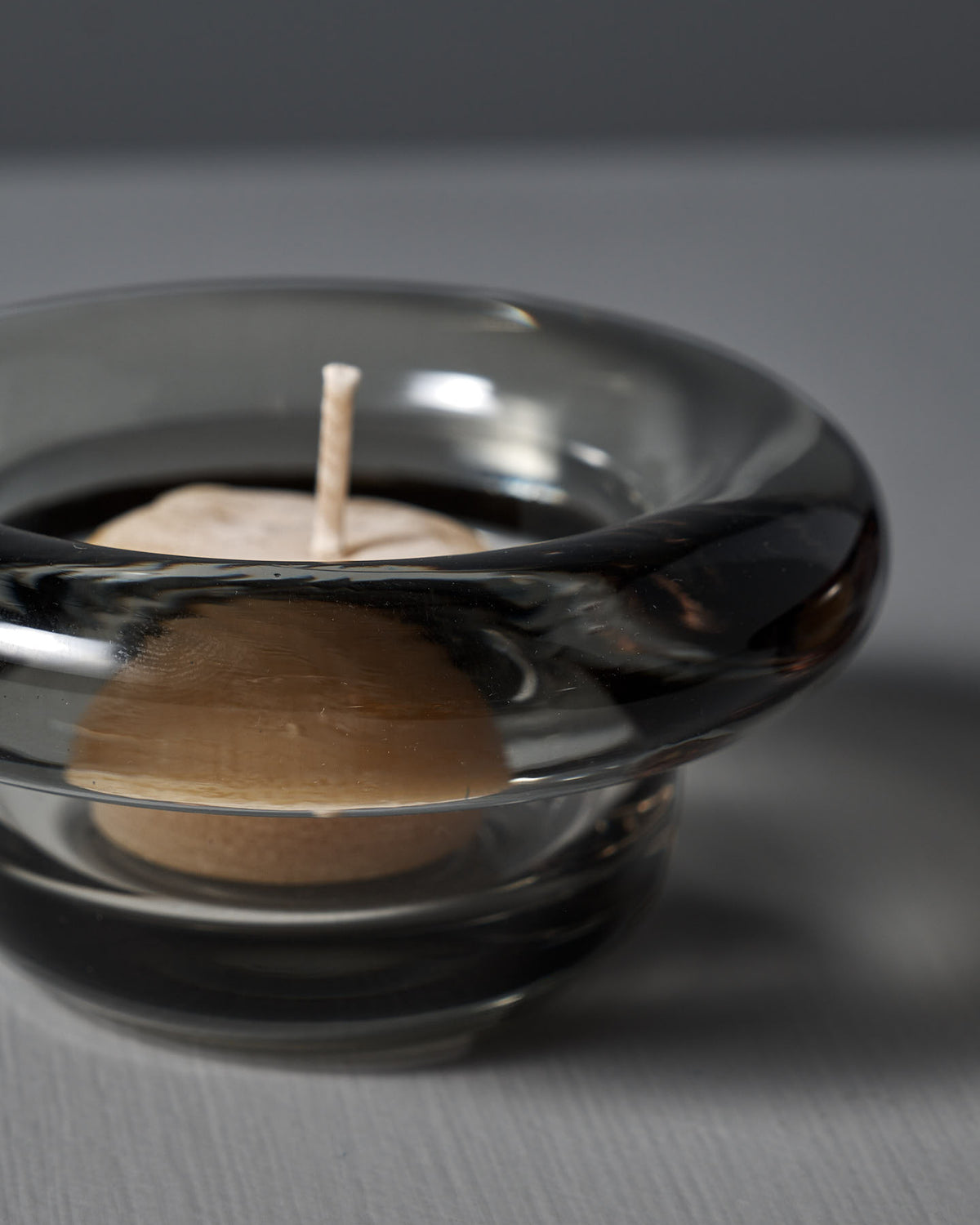 An unlit small candle is inside a Sirkel Candle Holder – Light Grey, crafted from pressed glass by Nedre Foss, on a gray surface.