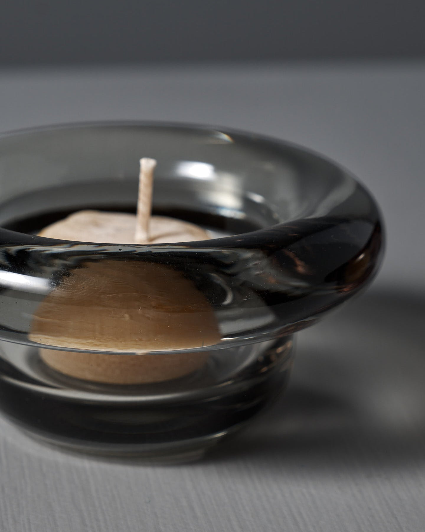 The Sirkel Candle Holder in translucent dark pressed glass by Nedre Foss features a wide, rounded rim and is elegantly suited as a tea-light holder on the smooth gray surface, enhancing the minimalist aesthetic.