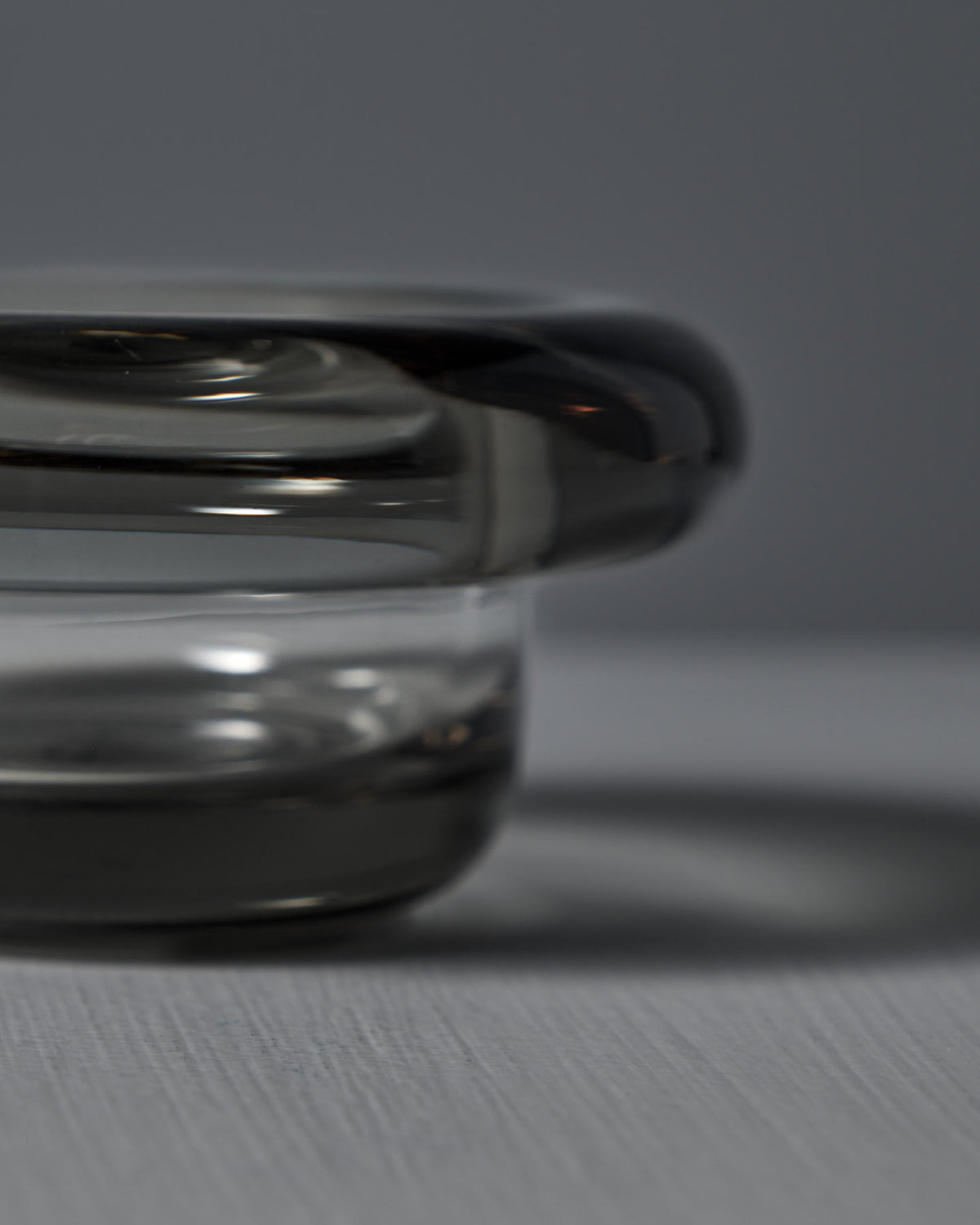 A close-up of the Sirkel Candle Holder by Nedre Foss shows its translucent, cylindrical design with a thick rim crafted from pressed dark glass, casting a shadow on a flat surface. The background is neutral gray.
