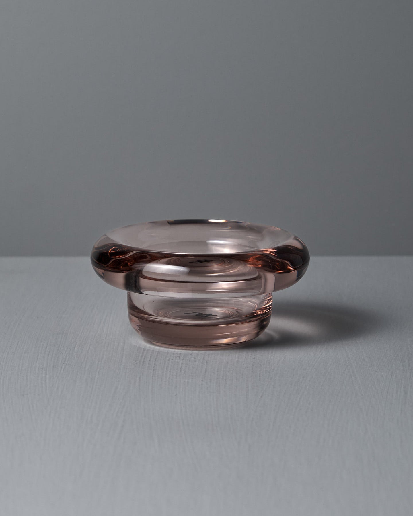 The Sirkel Candle Holder in Primrose Pink by Nedre Foss features a wide, rounded rim and sits on a textured surface against a gray background. Made from pressed glass with a subtle pink tint, it can also elegantly hold tea-lights.