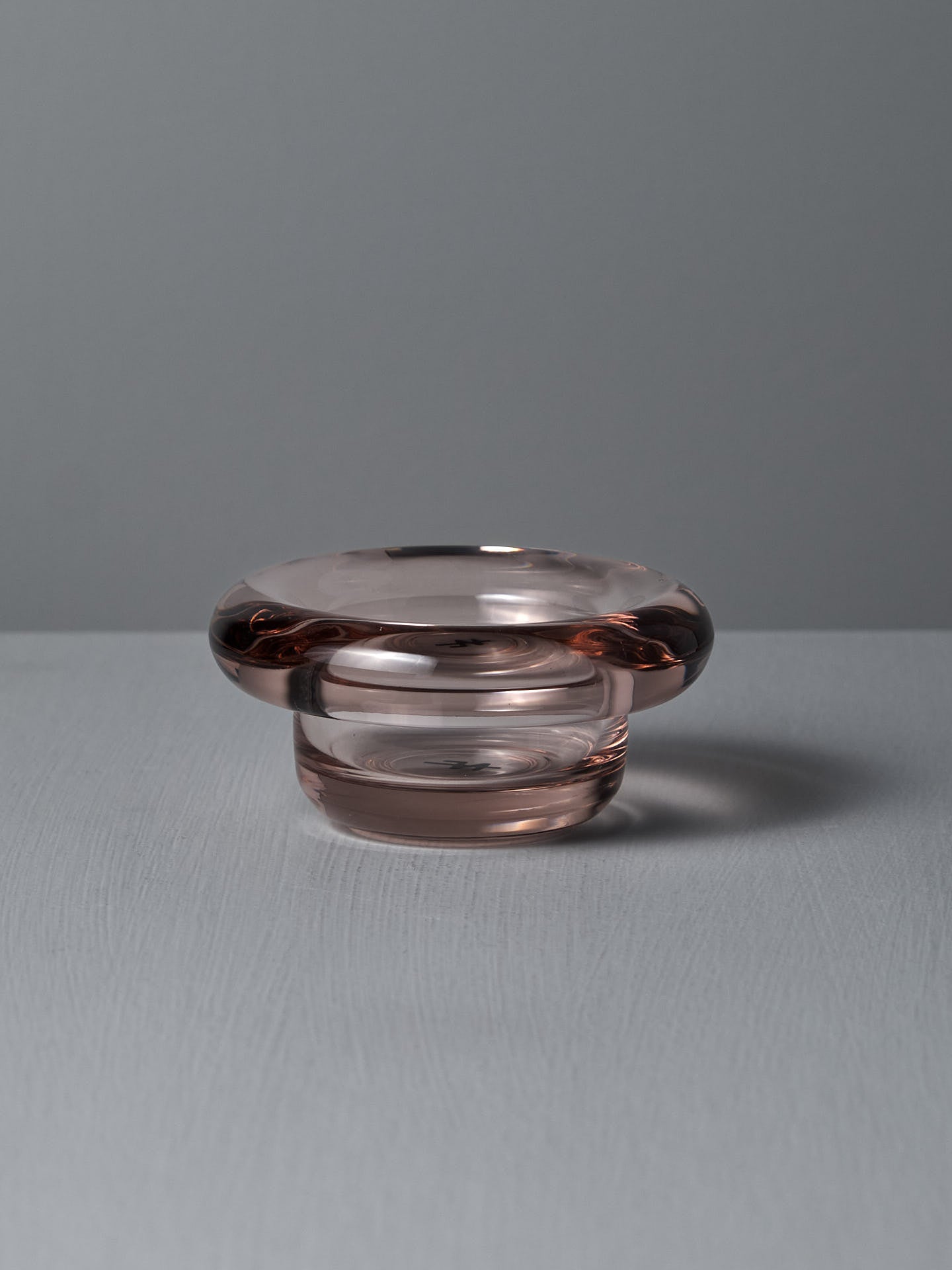 The Sirkel Candle Holder in Primrose Pink by Nedre Foss features a wide, rounded rim and sits on a textured surface against a gray background. Made from pressed glass with a subtle pink tint, it can also elegantly hold tea-lights.