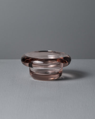 The Sirkel Candle Holder in Primrose Pink by Nedre Foss features a wide, rounded rim and sits on a textured surface against a gray background. Made from pressed glass with a subtle pink tint, it can also elegantly hold tea-lights.