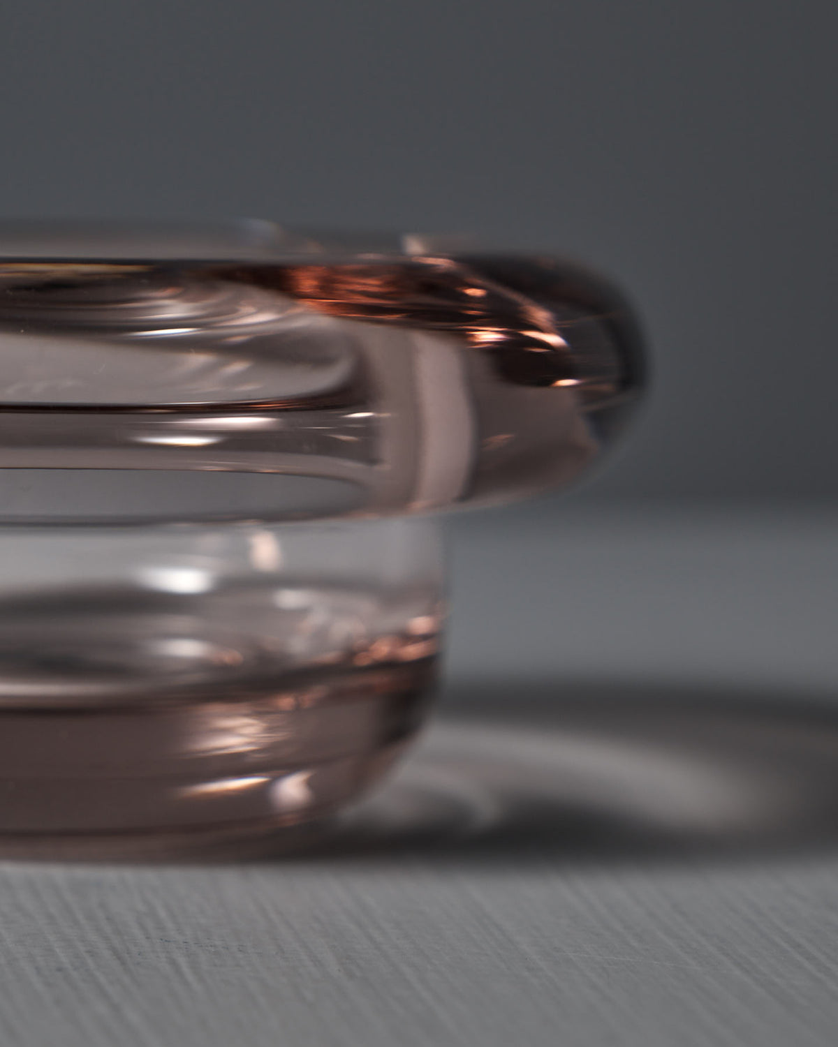 A close-up of the Nedre Foss Sirkel Candle Holder in Primrose Pink shows its transparent, rounded design casting a shadow on a light surface. The pressed glass has a slight pink tint, adding elegance to its simplicity.