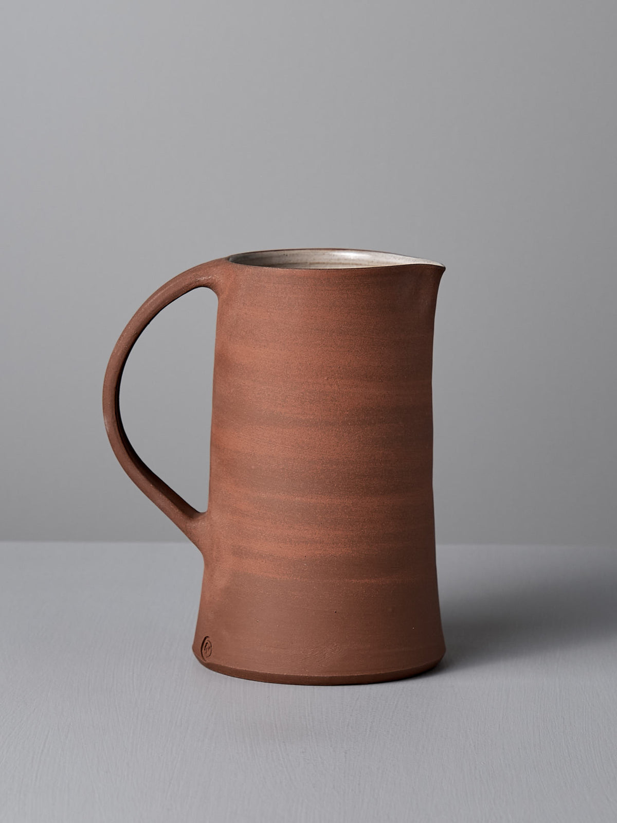 A handmade ceramic jug named &quot;Jug – Brick Red&quot; by Nicola Shuttleworth, featuring a handle and finished in satin glaze, stands on a grey surface against a light grey background.