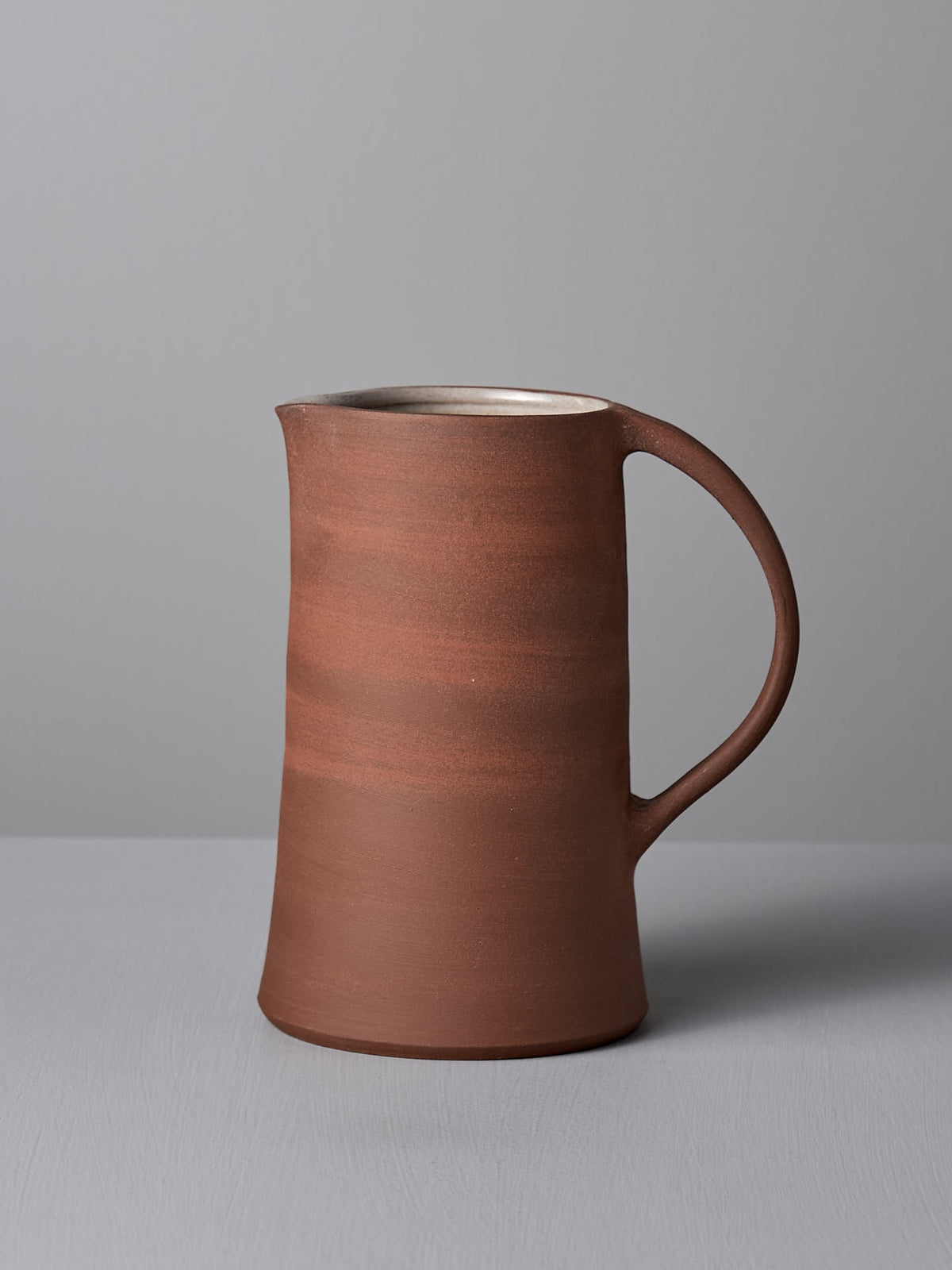 A brick red, handmade ceramic jug with a handle, from Nicola Shuttleworth&#39;s collection, placed on a light gray surface against a light gray background, showcases its charm beautifully.