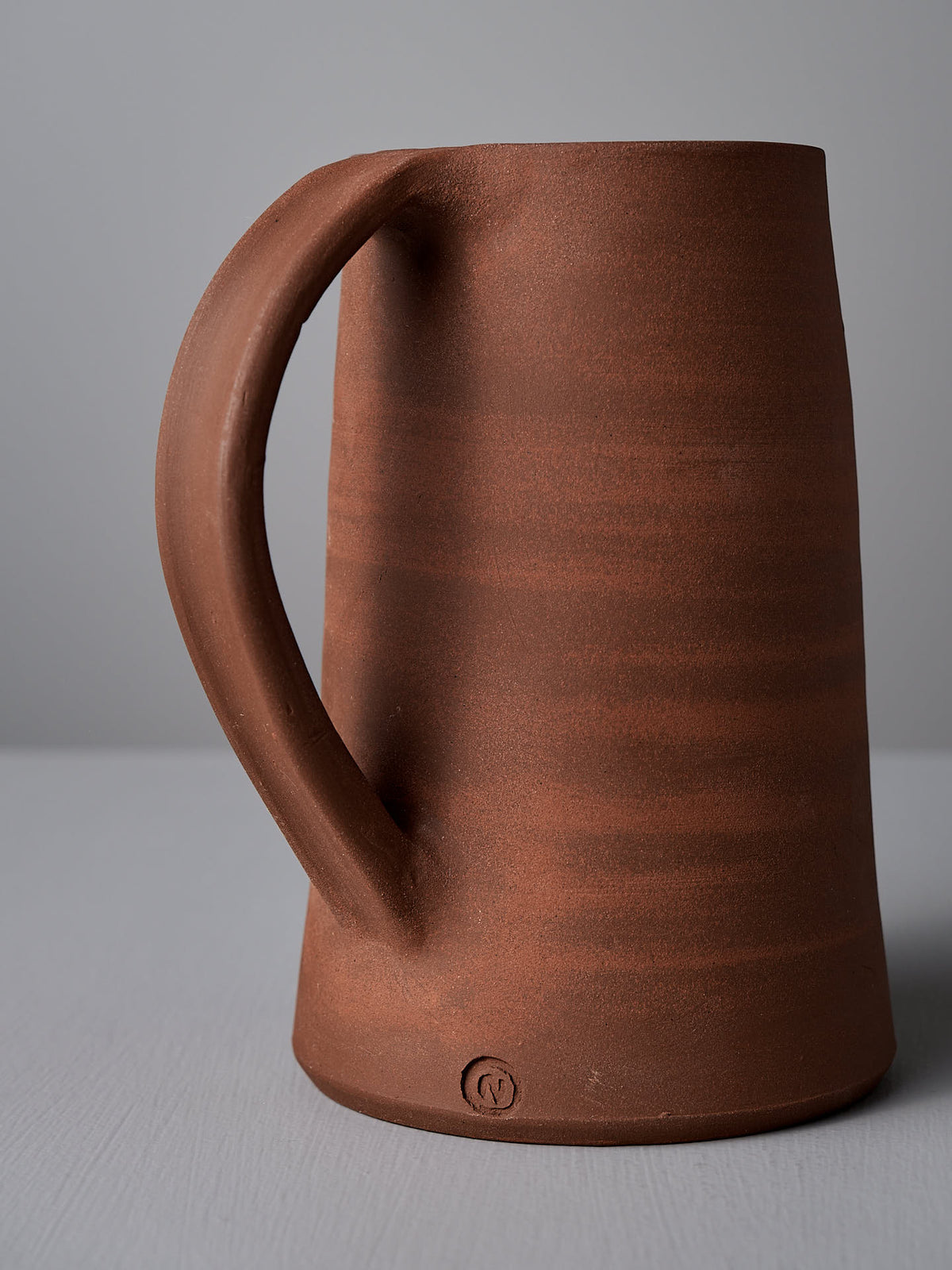 A brick red ceramic jug by Nicola Shuttleworth with a satin glaze and a large handle, set against a plain background.