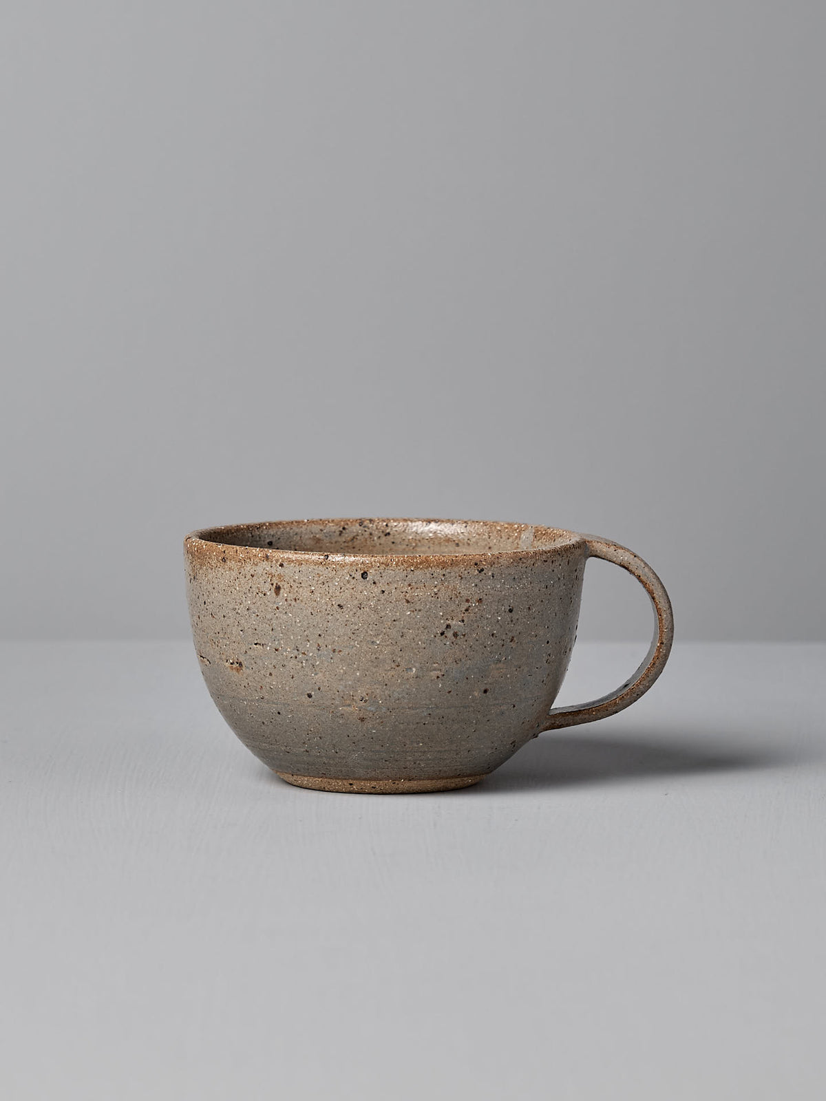 The &quot;Cup – Blue Ash&quot; by Nicola Shuttleworth, a speckled grey ceramic cup with a wide rim and handle, coated in a warm white satin glaze, rests on a grey surface against a light grey background.