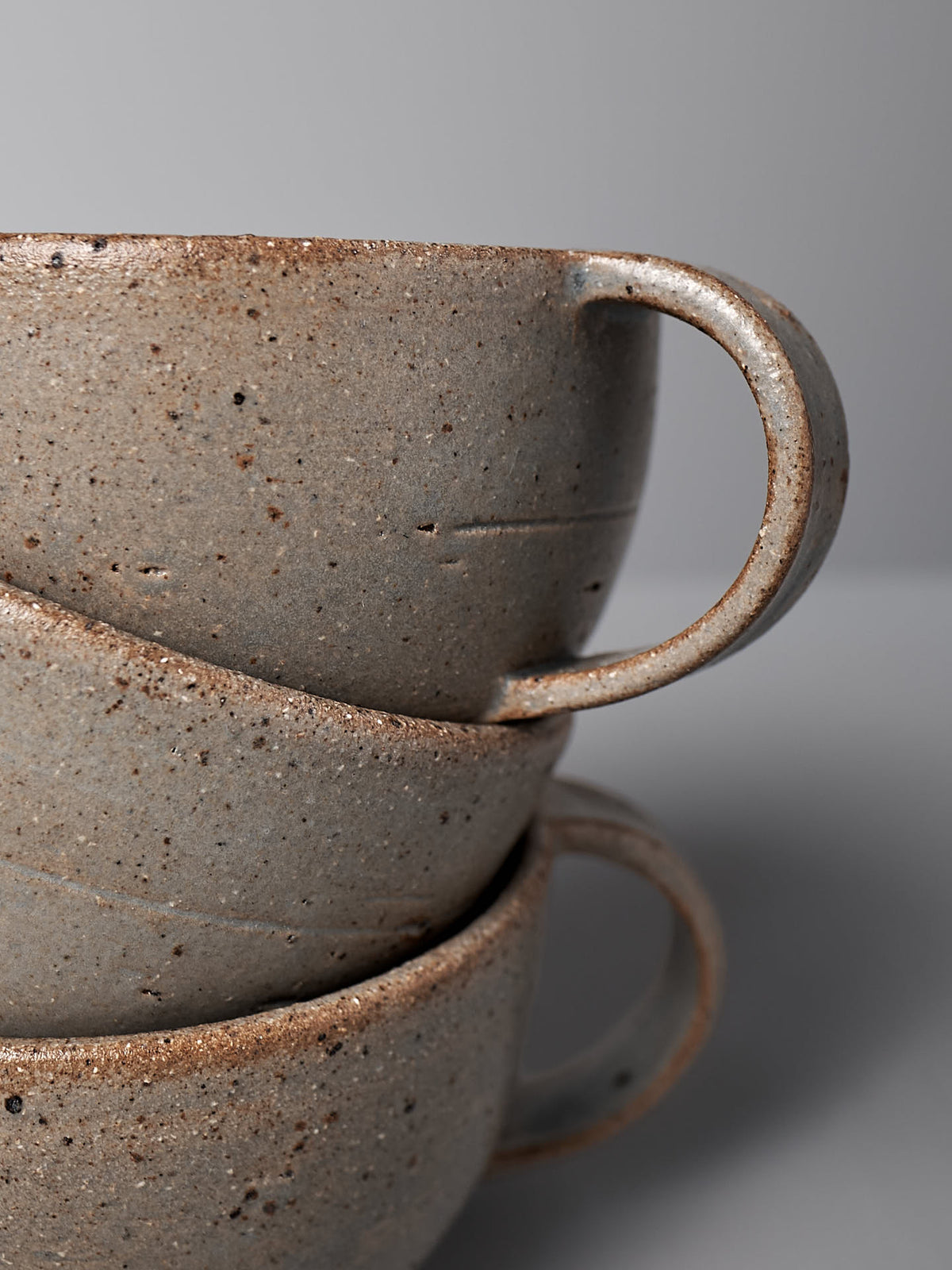 A stack of three blue ash stoneware cups by Nicola Shuttleworth, featuring handles and a minimalist design, positioned against a plain grey background.