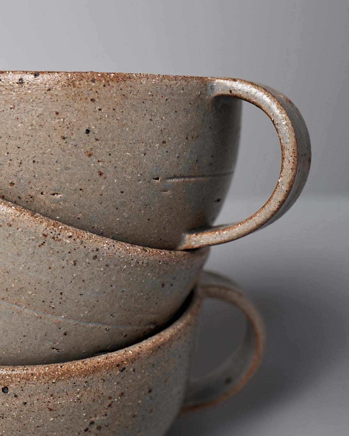 A stack of three blue ash stoneware cups by Nicola Shuttleworth, featuring handles and a minimalist design, positioned against a plain grey background.