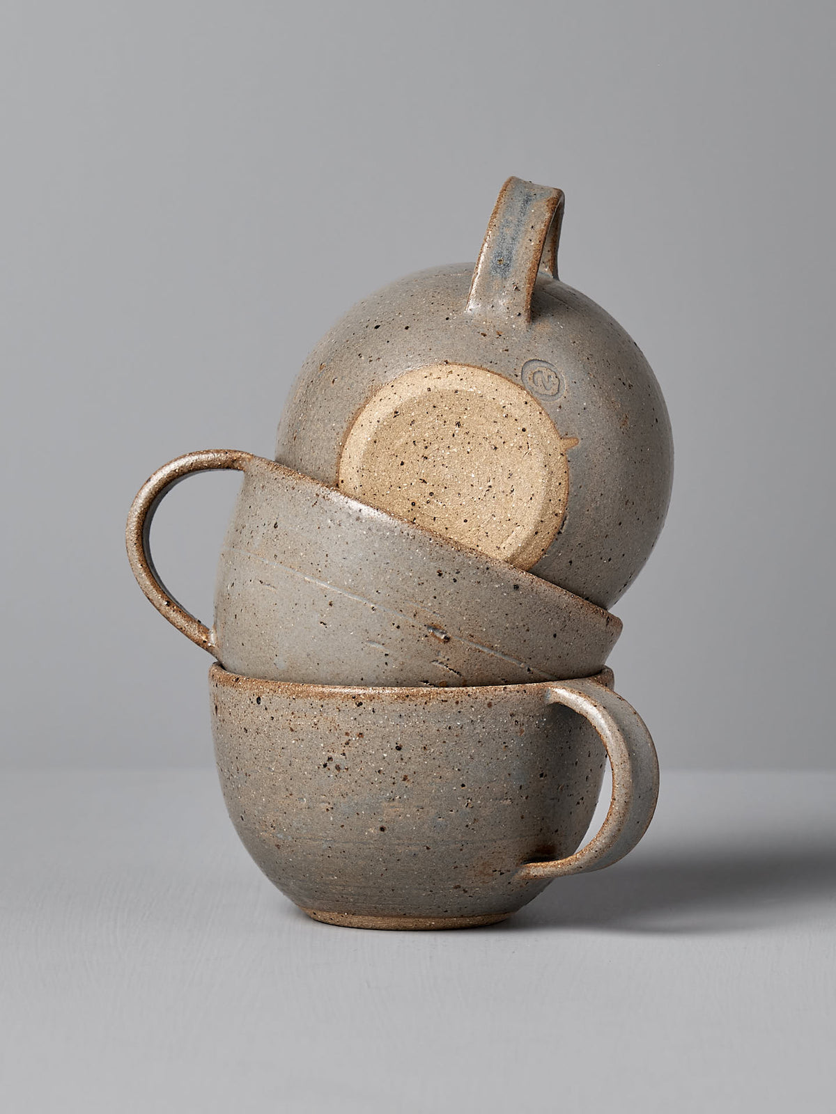 Three handmade ceramic cups, each from Nicola Shuttleworth&#39;s &quot;Cup – Blue Ash&quot; collection, featuring a warm white satin glaze and wide rims, stacked on top of each other against a plain gray background.