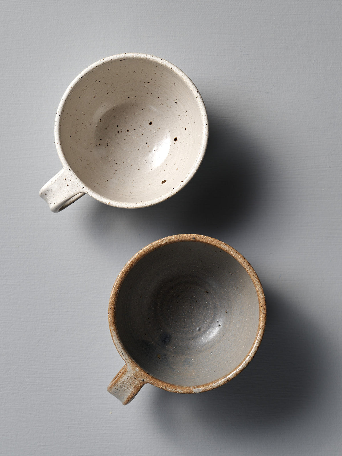 Two &quot;Cup – Speckled White&quot; ceramic cups, one light beige and one dark gray, sit side by side on a plain gray surface, each with a smooth satin glaze.