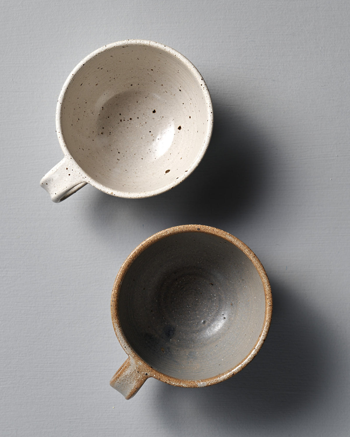 Two "Cup – Speckled White" ceramic cups, one light beige and one dark gray, sit side by side on a plain gray surface, each with a smooth satin glaze.