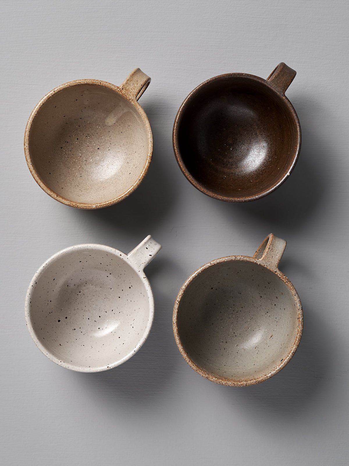 Four ceramic mugs in shades of beige, dark brown, white, and light gray are arranged in a 2x2 grid on a flat, light gray surface. Each wide-rimmed cup from the Nicola Shuttleworth collection has a handle and they are viewed from above. The speckled white cup stands out among them with its unique texture.