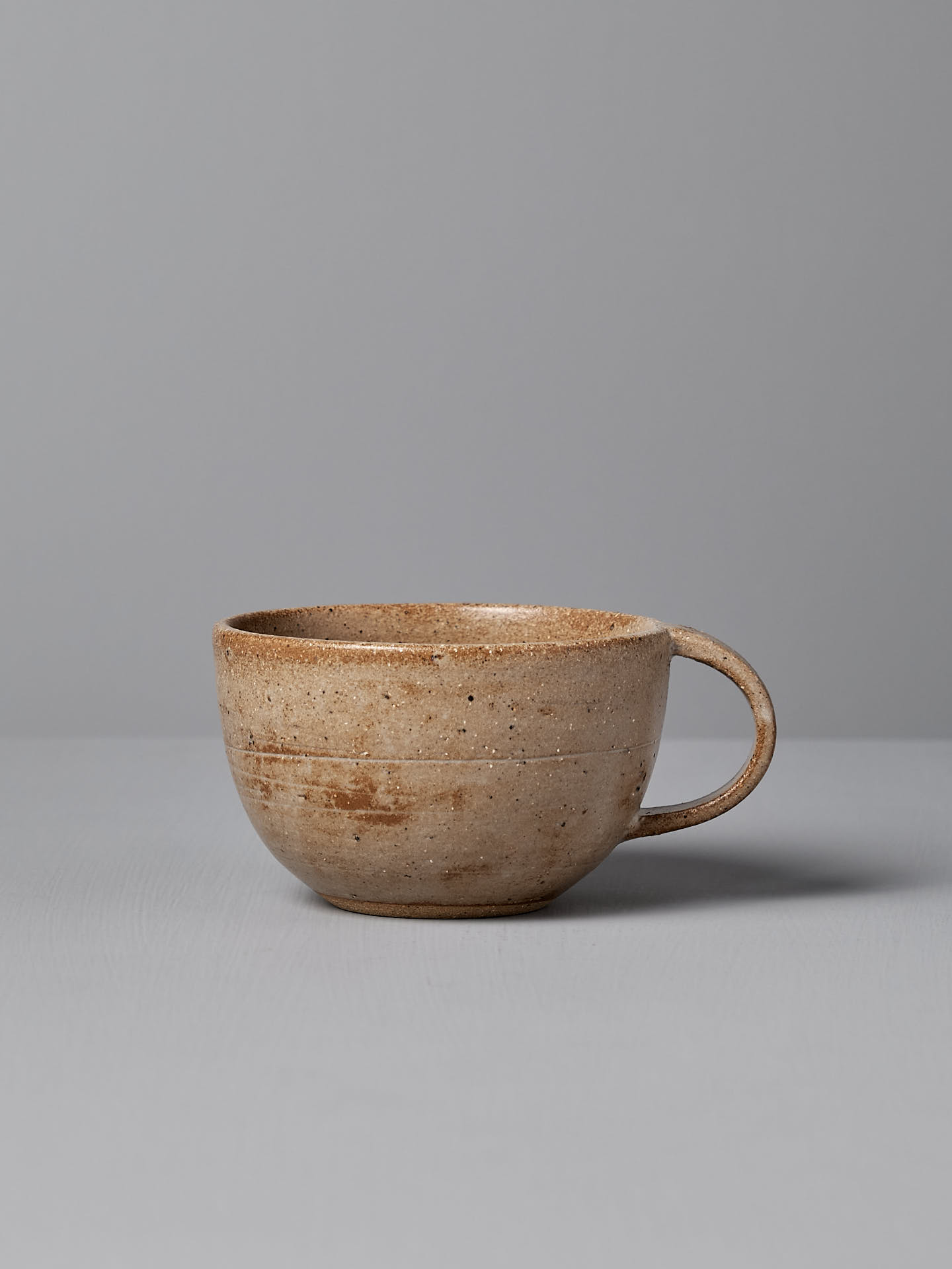 A Nicola Shuttleworth Cup – Light Brown, handmade with a handle and a wide rim, adorned with a blue ash satin glaze, sits on a gray surface against a gray background.