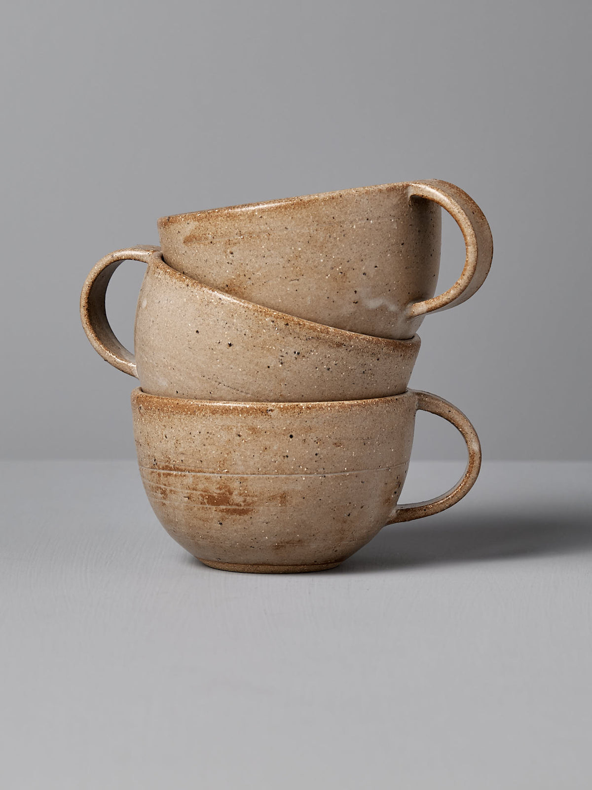 Three light brown stoneware coffee mugs, each featuring a stunning Nicola Shuttleworth design, are stacked on top of each other against a neutral grey background.