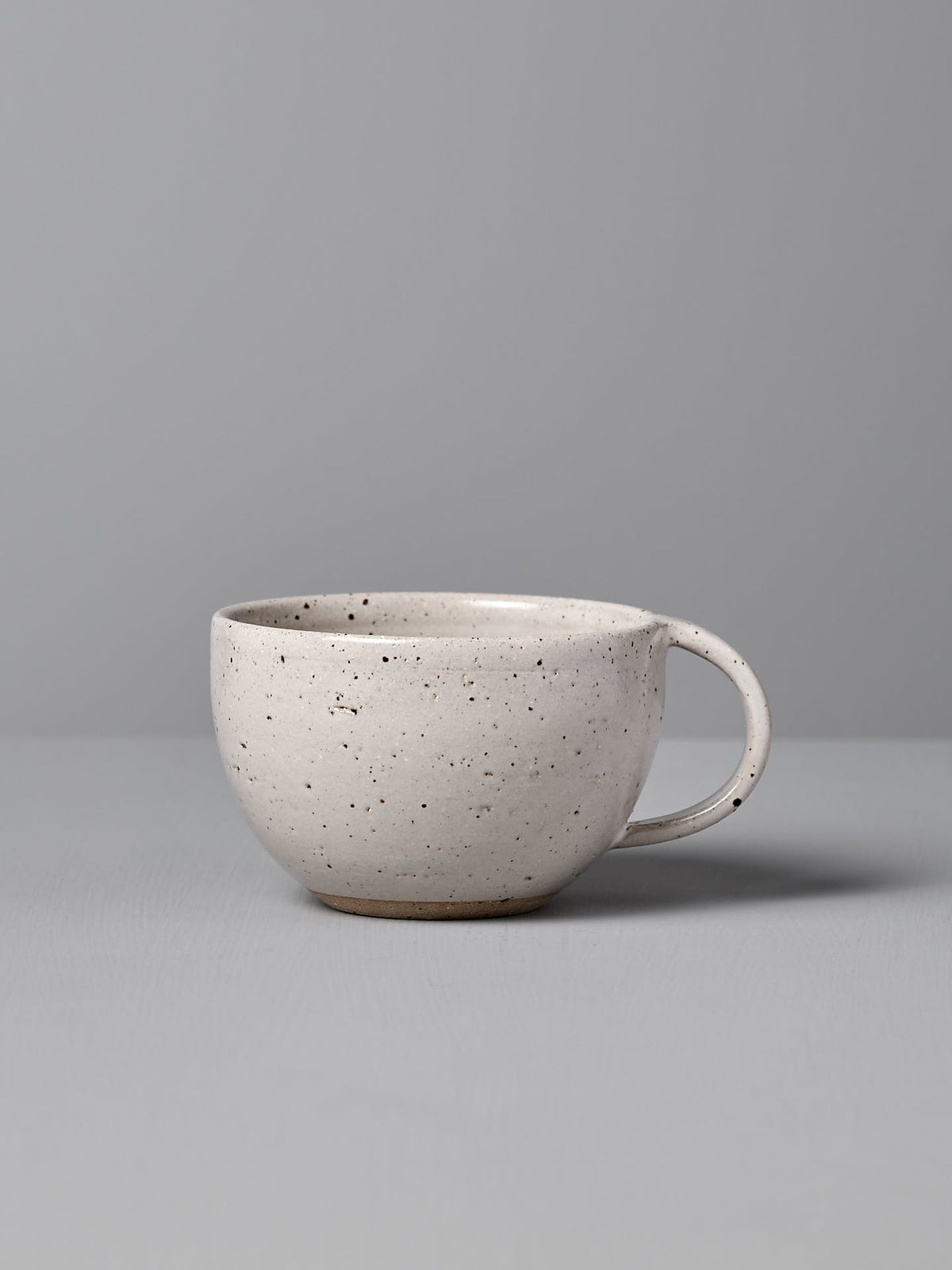 The Nicola Shuttleworth &quot;Cup – Speckled White,&quot; featuring a round handle and a warm white satin glaze, sits on a light gray surface against a light gray background.