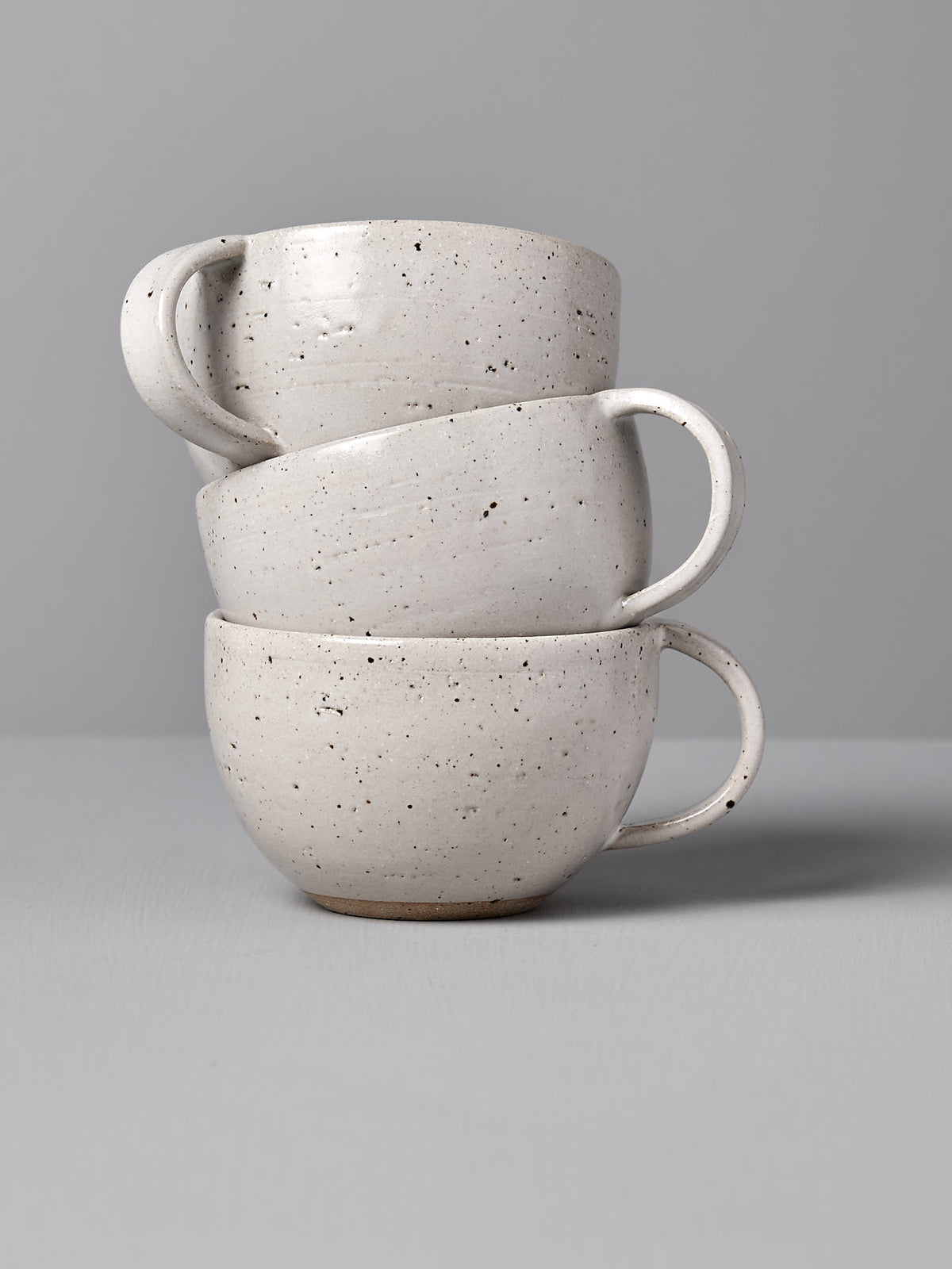 Three Nicola Shuttleworth &quot;Cup – Speckled White&quot; handmade ceramic cups are stacked on top of each other against a gray background, showcasing their speckled detailing and warm white satin glaze.