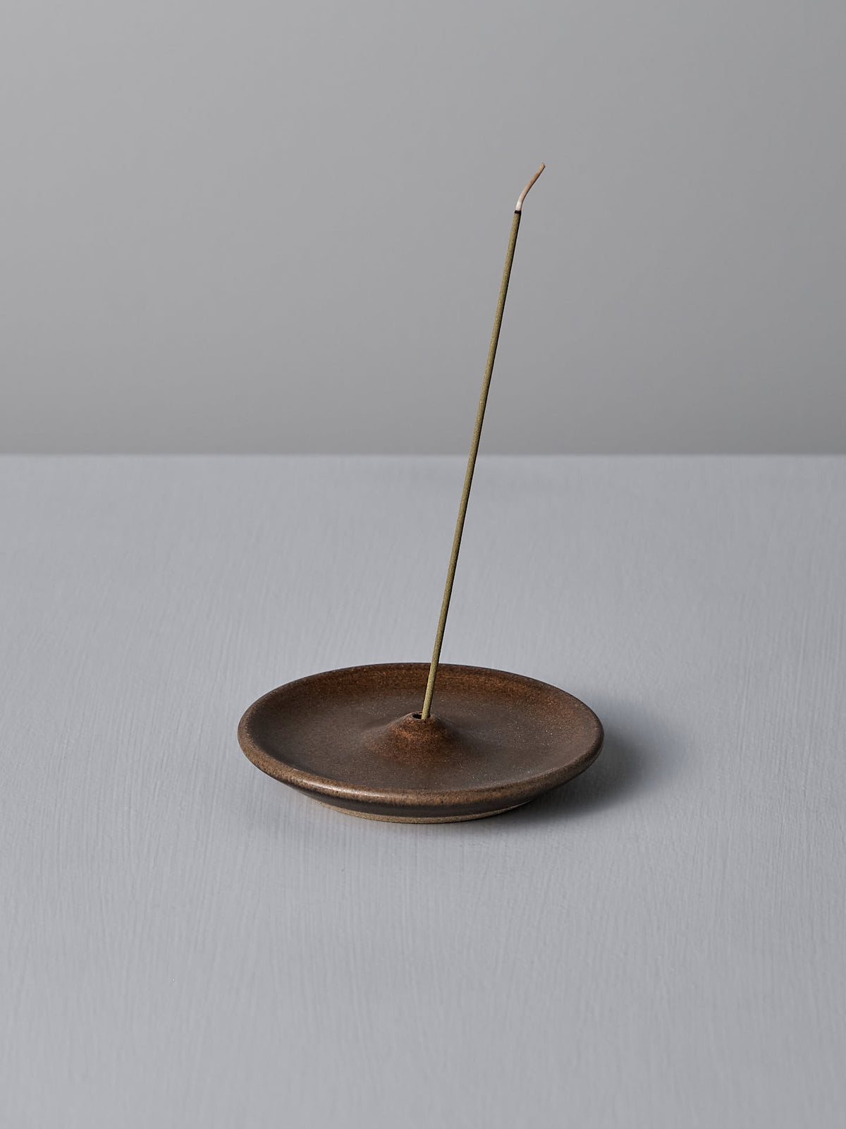 A thin incense stick stands upright in the Nicola Shuttleworth Incense Holder – Brown, a simple, handmade stoneware piece, on a light gray surface.