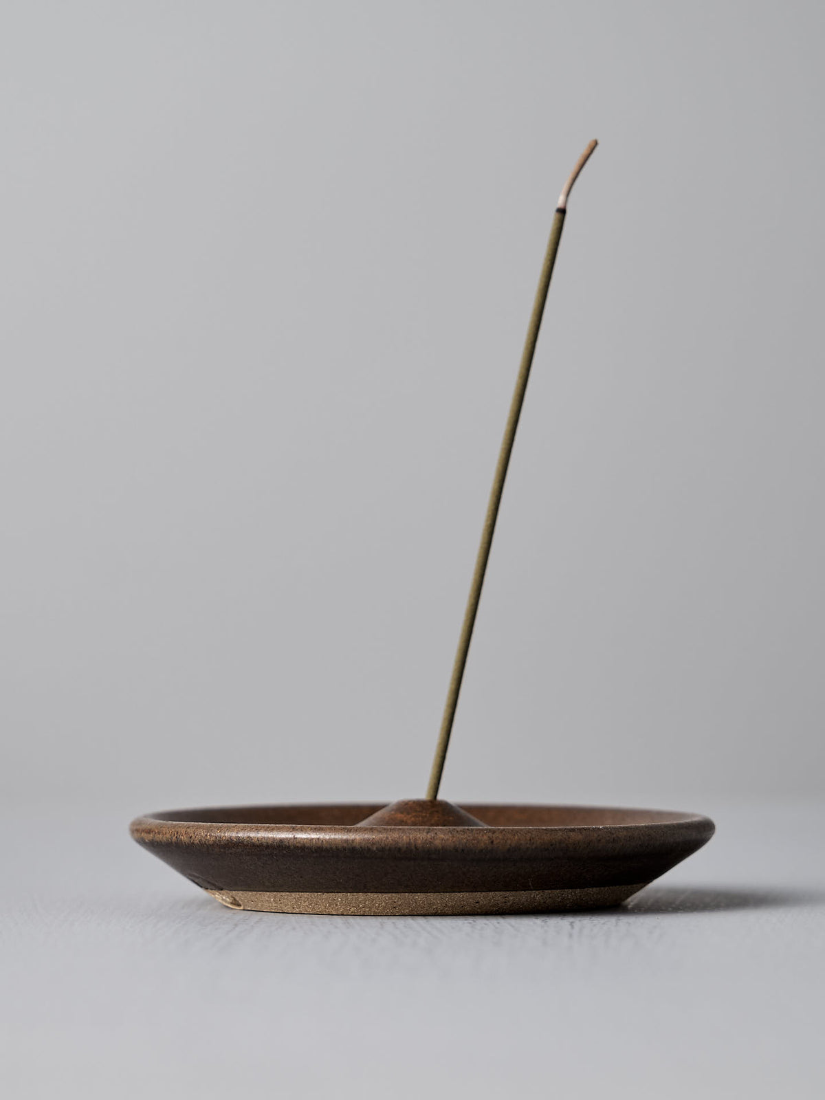 A single unlit incense stick stands upright in the Nicola Shuttleworth Incense Holder – Brown, a small, handmade stoneware piece against a plain background.