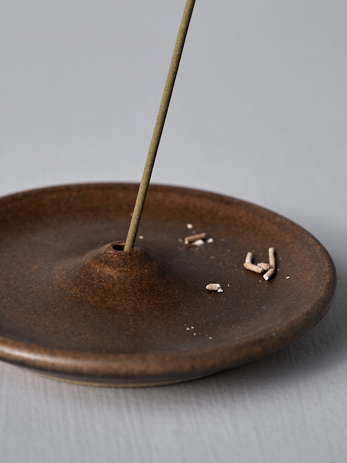 A lighted incense stick stands upright in a handmade Nicola Shuttleworth &quot;Incense Holder – Brown&quot; stoneware holder, with a small pile of ashes on the dish.