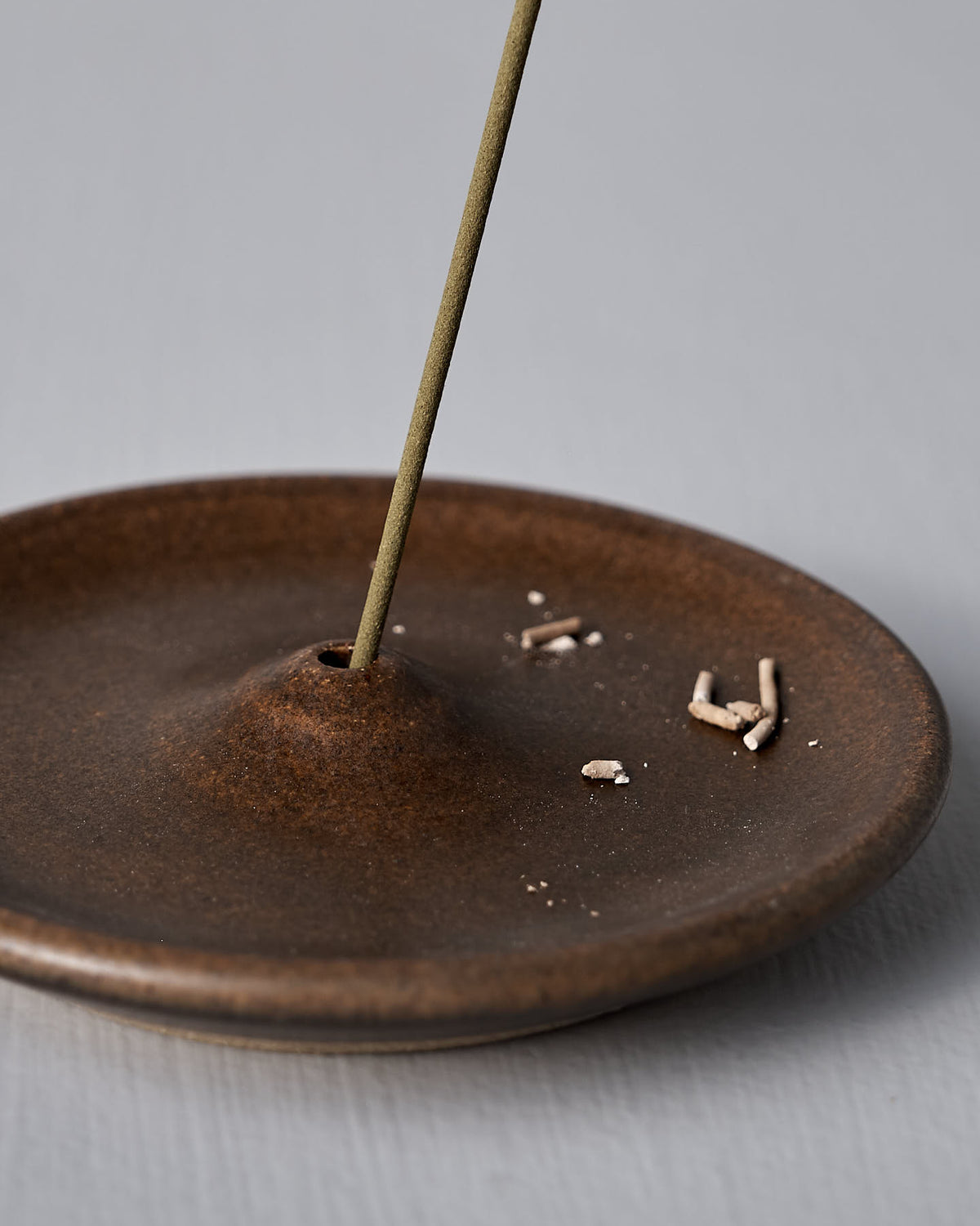 A lighted incense stick stands upright in a handmade Nicola Shuttleworth "Incense Holder – Brown" stoneware holder, with a small pile of ashes on the dish.