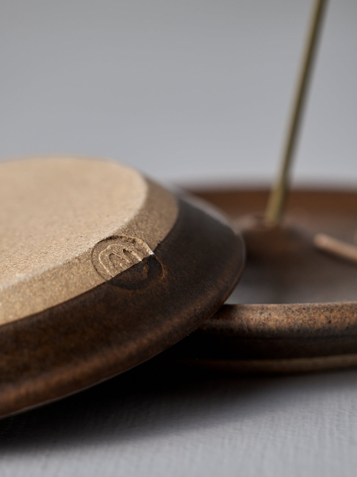 Close-up of Nicola Shuttleworth&#39;s &quot;Incense Holder – Brown,&quot; a handmade stoneware piece with a distinct two-tone glaze, resting on a metal rod. A small maker&#39;s mark is visible on the edge of the dish.