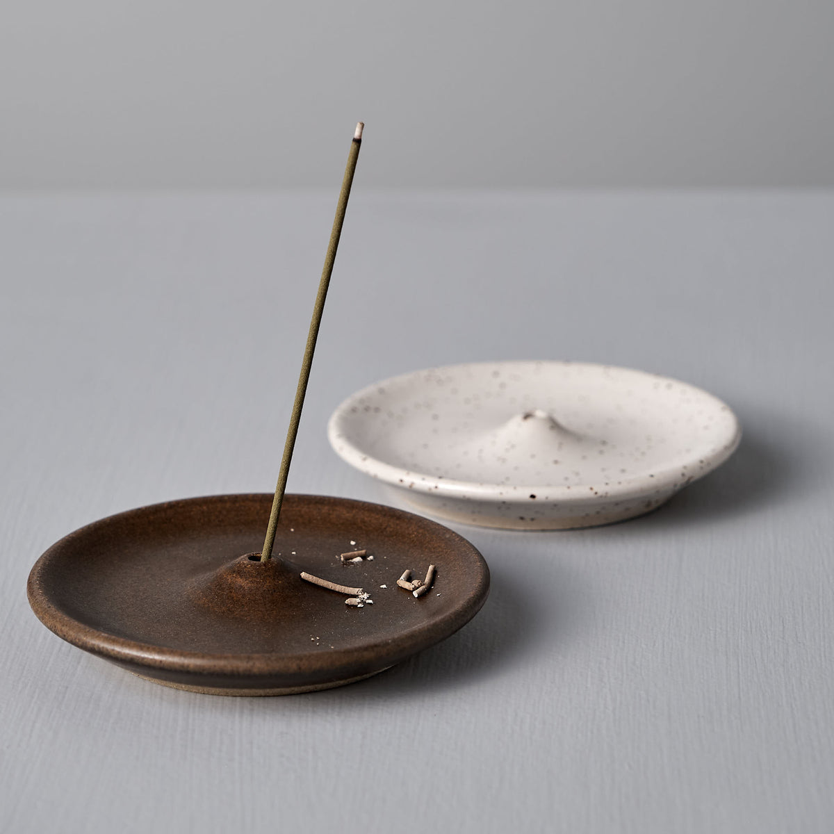 Two Wellington handcrafted stoneware incense holders, one from Nicola Shuttleworth&#39;s collection in a brown finish and another named the Speckled White Incense Holder, are placed on a gray surface. The brown holder contains a burning incense stick with small ash fragments, while the Speckled White Incense Holder is empty.