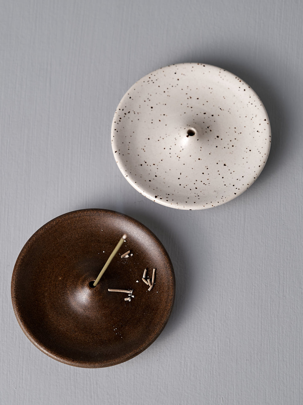 Two Nicola Shuttleworth handcrafted ceramic incense holders rest on a grey surface. The Incense Holder – Speckled White is empty, while the brown stoneware incense holder contains incense stubs and ash.