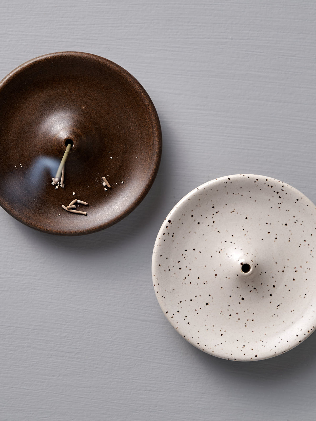 Placed on a light grey surface, a pair of round ceramic incense holders captures attention: one in a speckled white finish and the other in an earthy brown adorned with burnt incense sticks. These handmade stoneware pieces, known as the &quot;Incense Holder – Speckled White&quot; by Nicola Shuttleworth, exude rustic charm and showcase Wellington&#39;s handcrafted artistry.