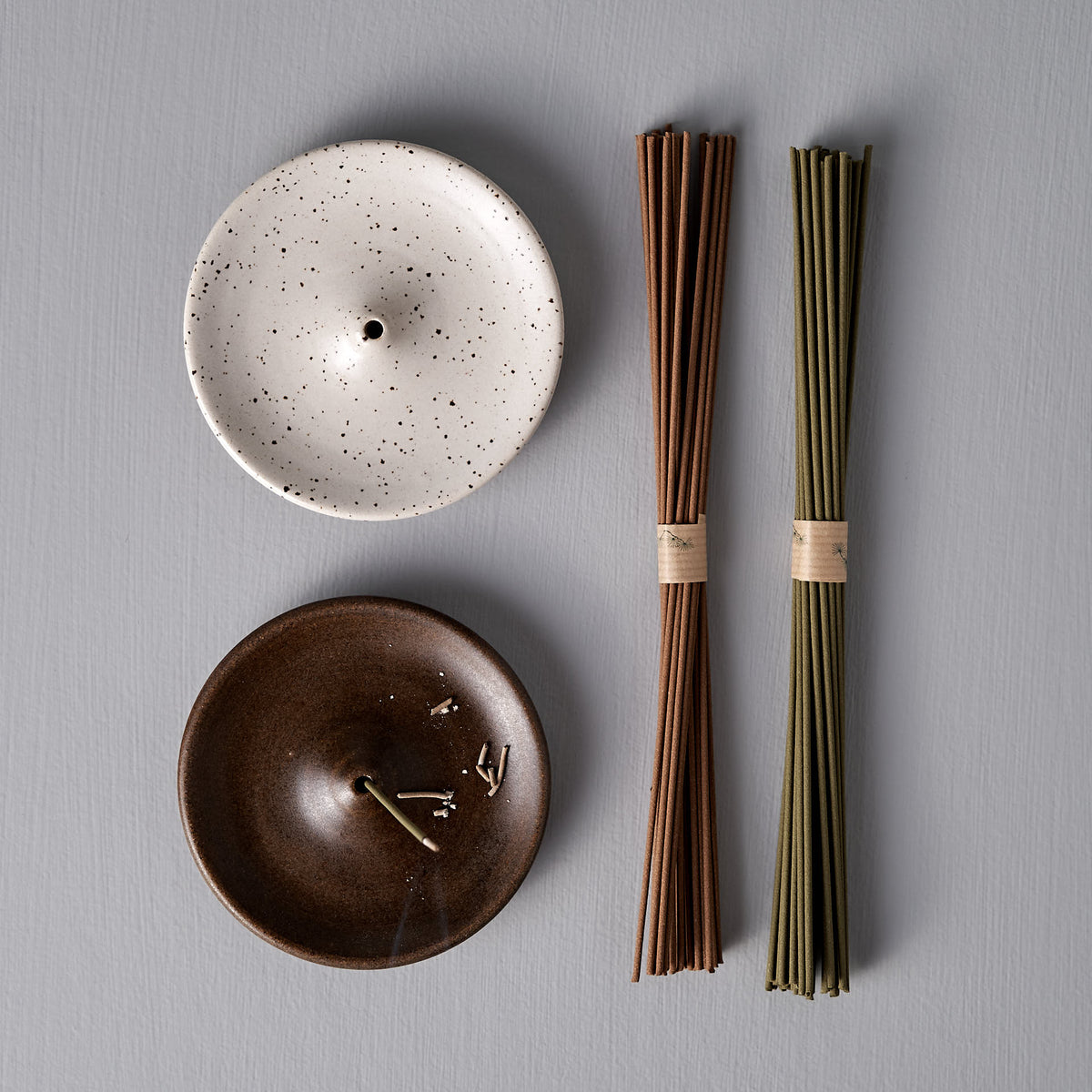 Two handmade Incense Holders – Speckled White by Nicola Shuttleworth and two bundles of incense sticks, one in brown and one in green, are carefully arranged on a flat gray surface. Crafted with artisanal elegance, these pieces enhance your space with their sophisticated charm.