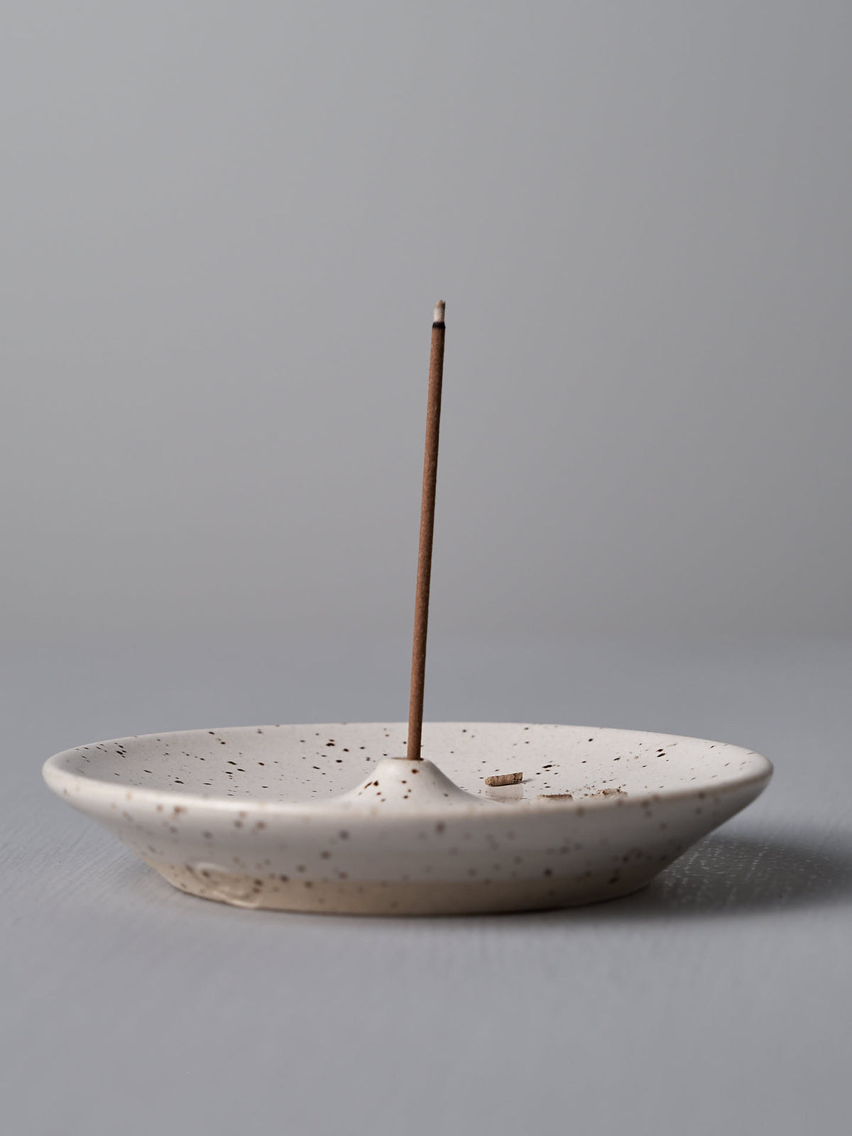 A single unlit incense stick stands upright in a Nicola Shuttleworth Incense Holder – Speckled White handcrafted stoneware against a plain background.