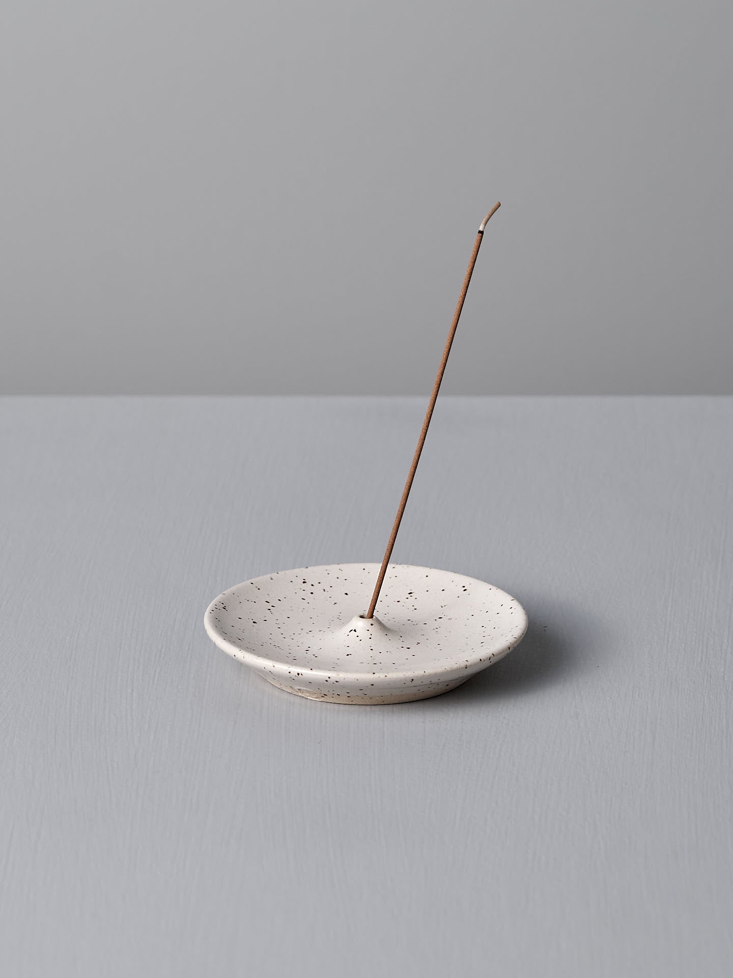 A lit incense stick standing upright in the Nicola Shuttleworth Incense Holder – Speckled White on a gray surface.